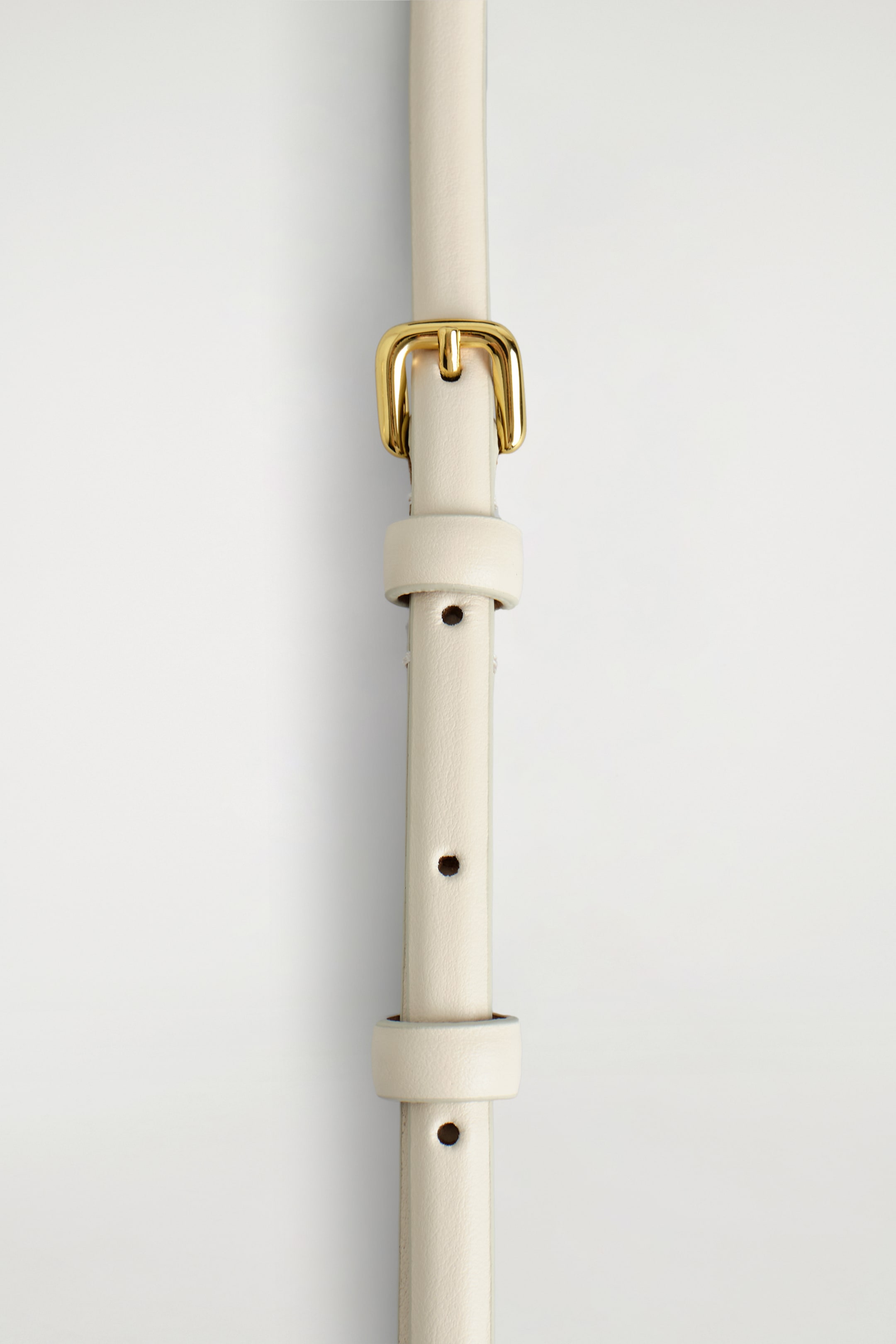 Agrandir l'image: Une ceinture en cuir mince de couleur crème est disposée verticalement, affichant une boucle classique dorée en haut. La sangle de la ceinture présente plusieurs trous d'ajustement ronds et deux passants en cuir assortis.