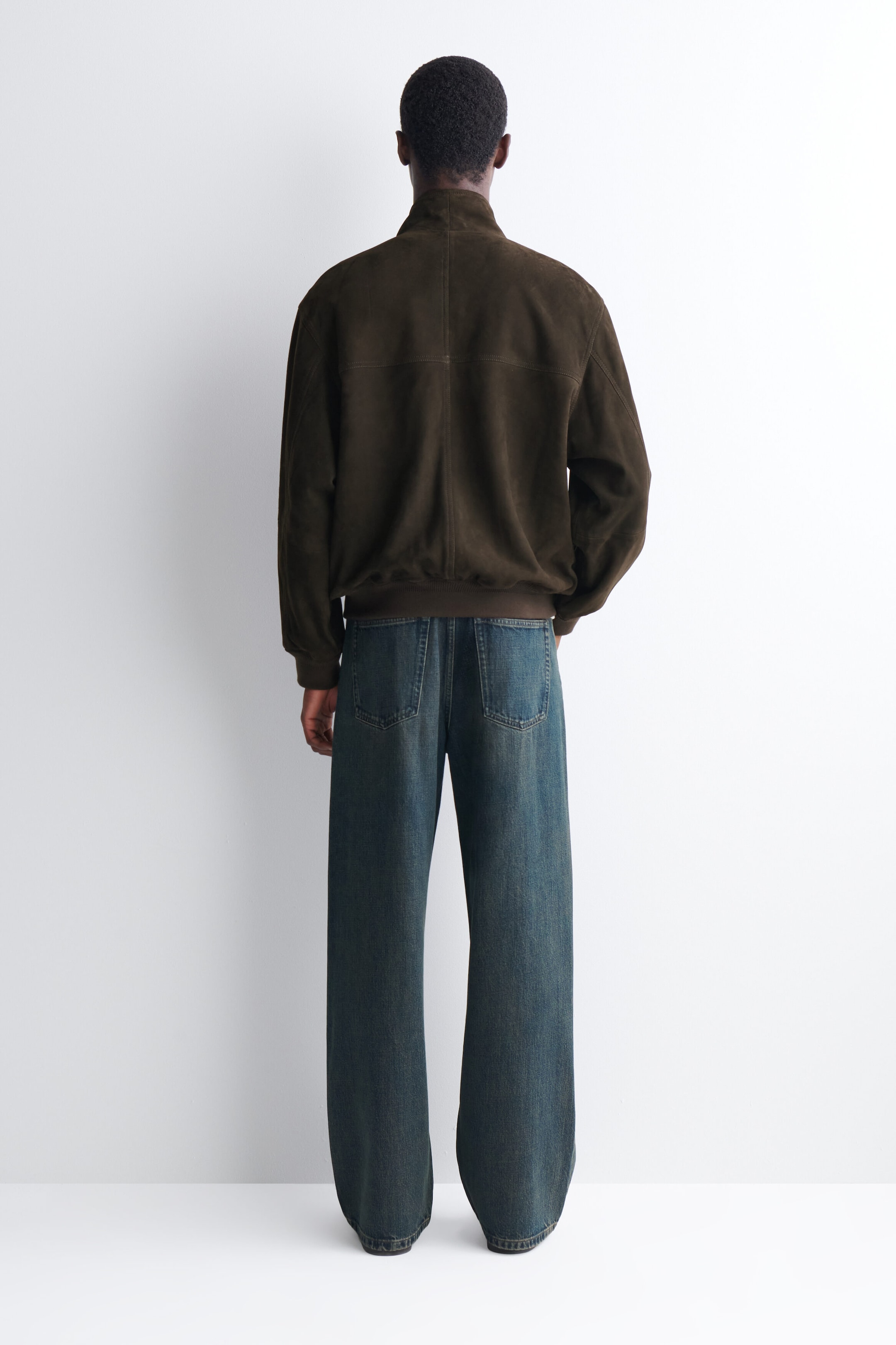 A dark brown suede bomber jacket with a high collar and ribbed hem is being worn by the model, who is shown from behind. The jacket is paired with wide-leg, mid-wash blue jeans. A dark brown suede bomber jacket with a high collar and ribbed hem is being worn by the model, who is shown from behind. The jacket is paired with wide-leg, mid-wash blue jeans.