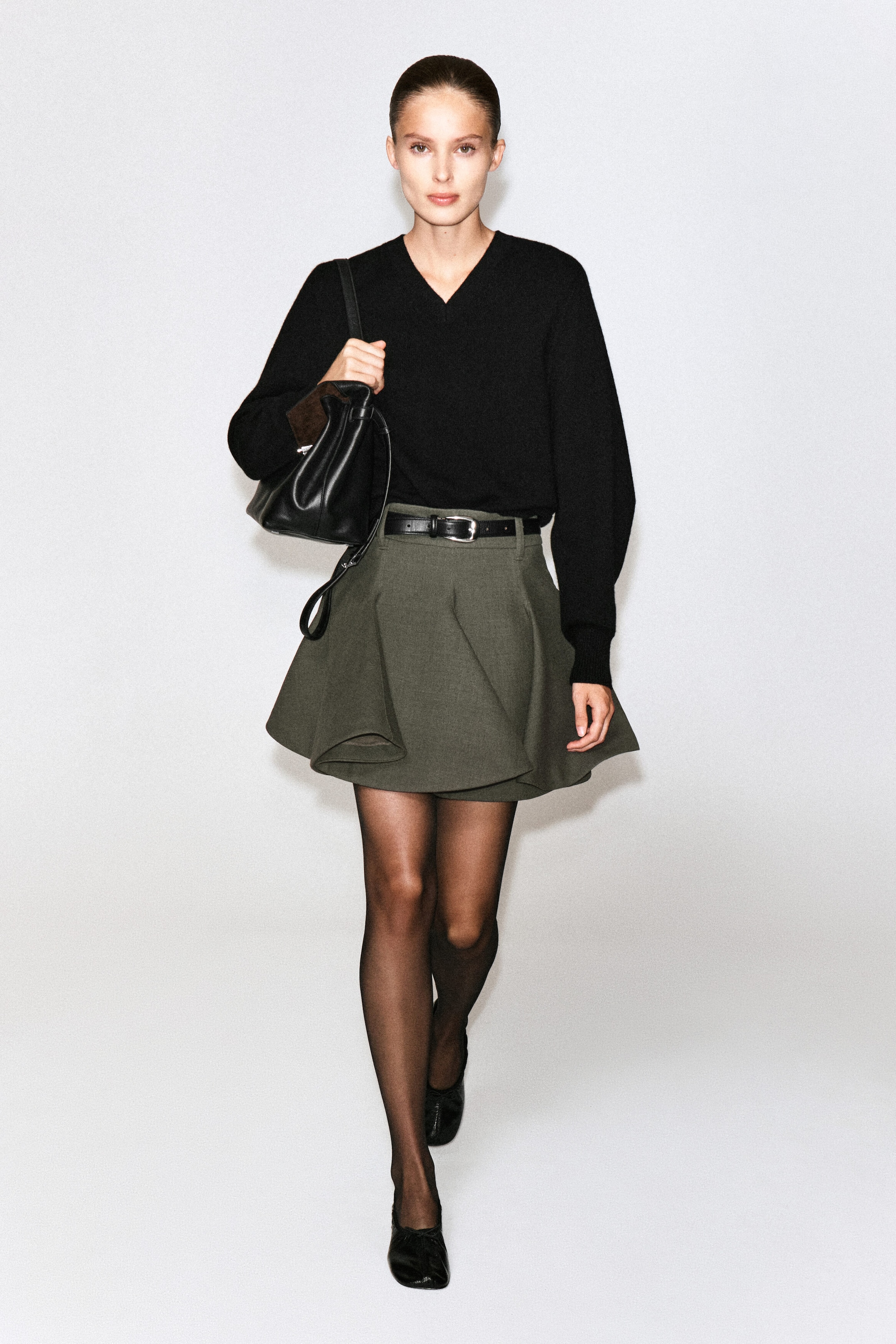 This sculptural tailored wool mini skirt in dark olive green is worn by the model, showcasing a flared, structured silhouette with draped folds. The skirt is styled with a black leather belt at the waist and paired with a black V-neck sweater, sheer black tights, and black flats. This sculptural tailored wool mini skirt in dark olive green is worn by the model, showcasing a flared, structured silhouette with draped folds. The skirt is styled with a black leather belt at the waist and paired with a black V-neck sweater, sheer black tights, and black flats.
