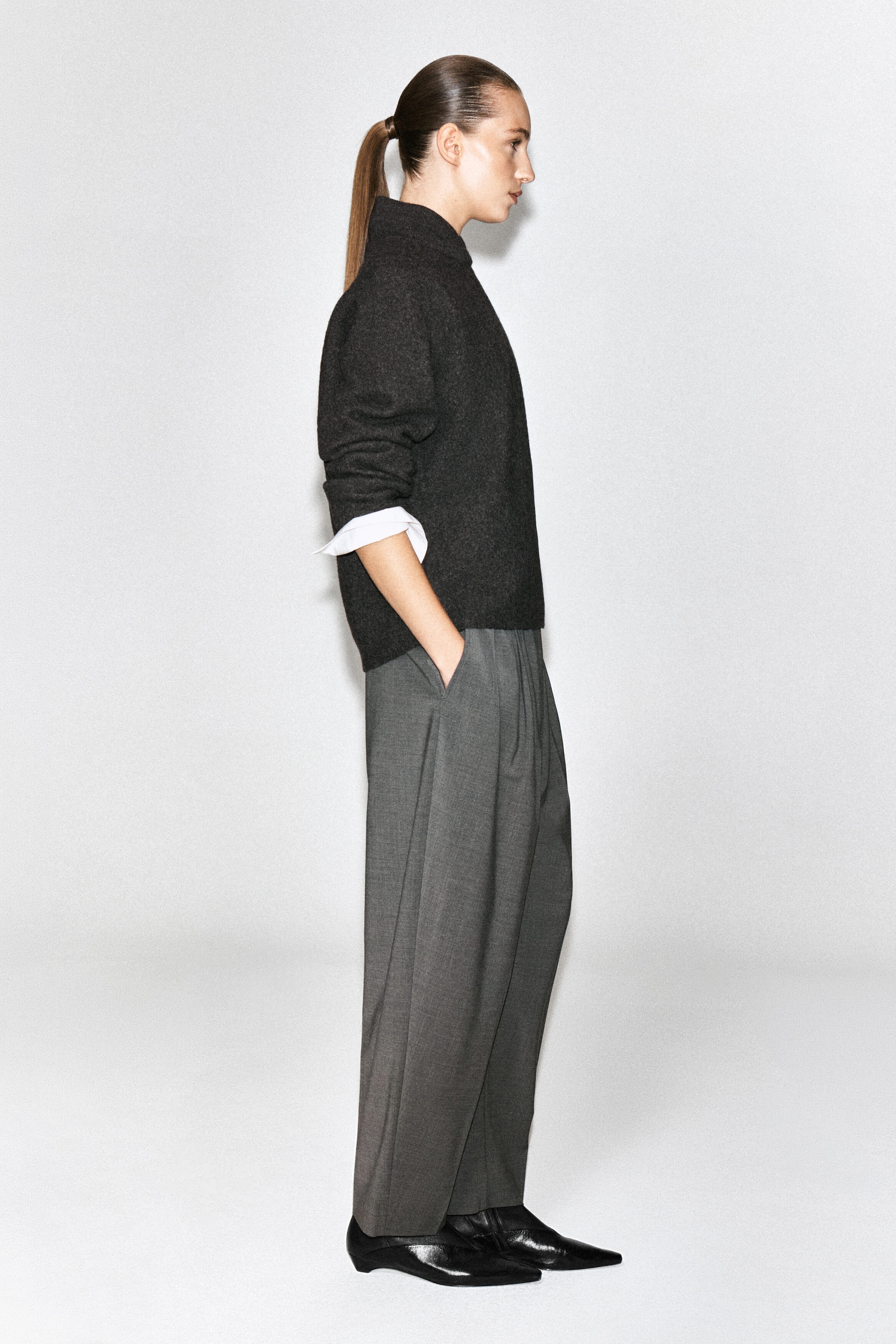 A layered-collar boiled wool sweater in dark gray is being worn by the model, featuring a high, structured neck and long sleeves. The sweater is styled over a white shirt with visible cuffs and paired with wide-leg gray trousers.
