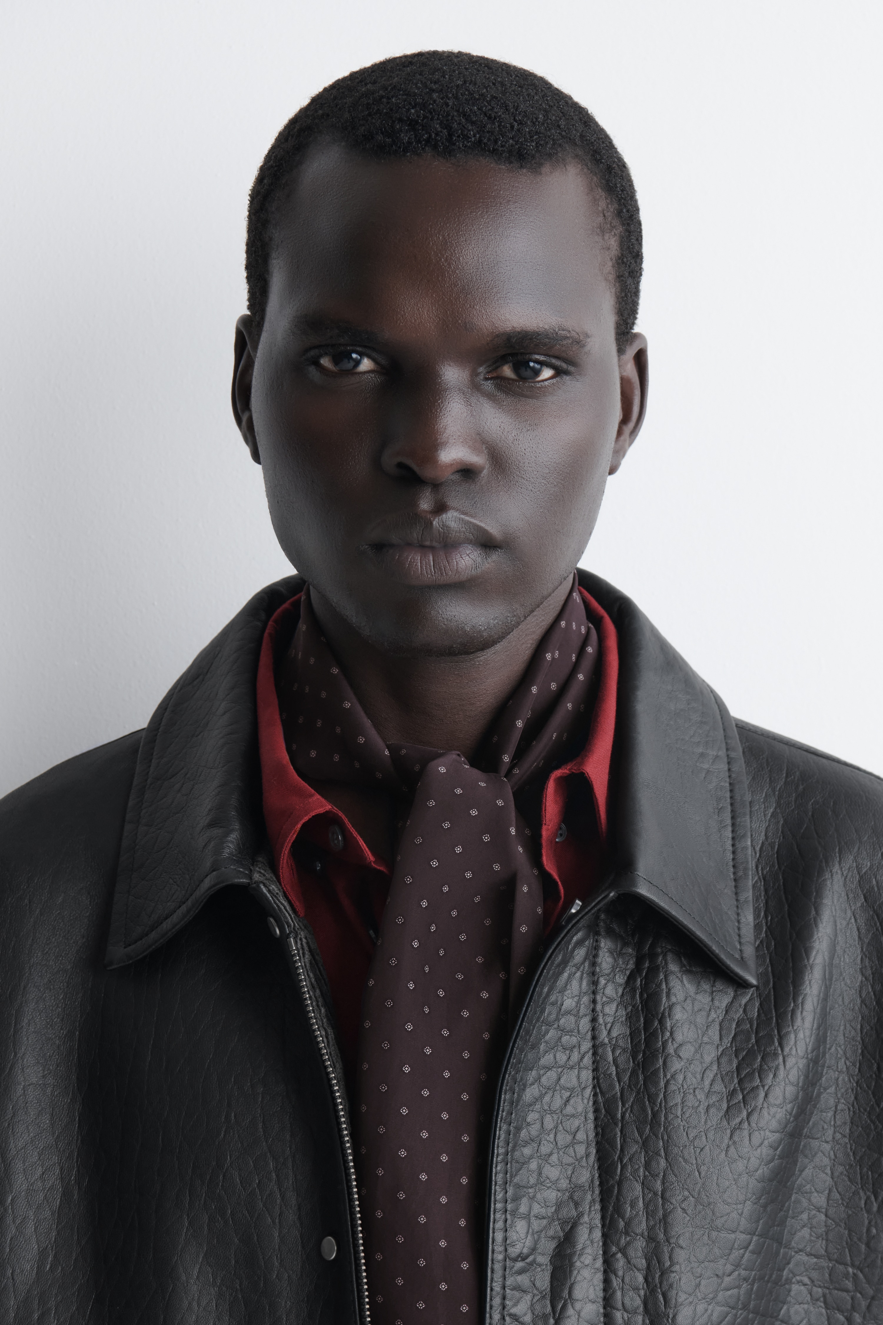 A slim, elongated dark brown silk scarf is hanging folded in half, displaying a repeating white micro-diamond motif across its body. The bottom sections feature a denser geometric diamond pattern above horizontal olive-green and white stripes, with solid dark brown ends.