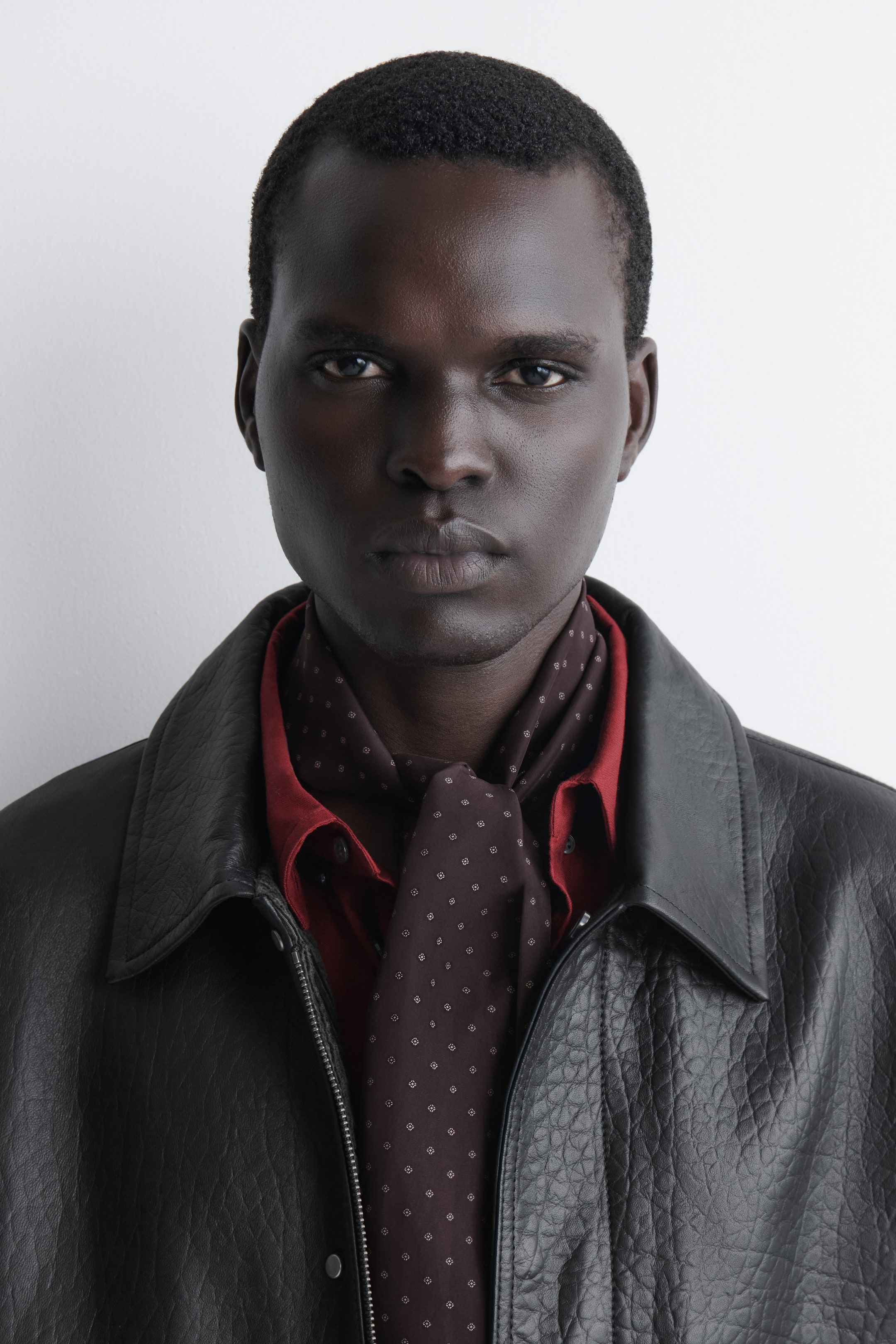 View larger image: A slim, dark brown silk scarf featuring a white micro-diamond motif is shown from the front, tied around the neck and draping down the chest. It is styled layered over a dark red collared shirt and under a textured black leather jacket.