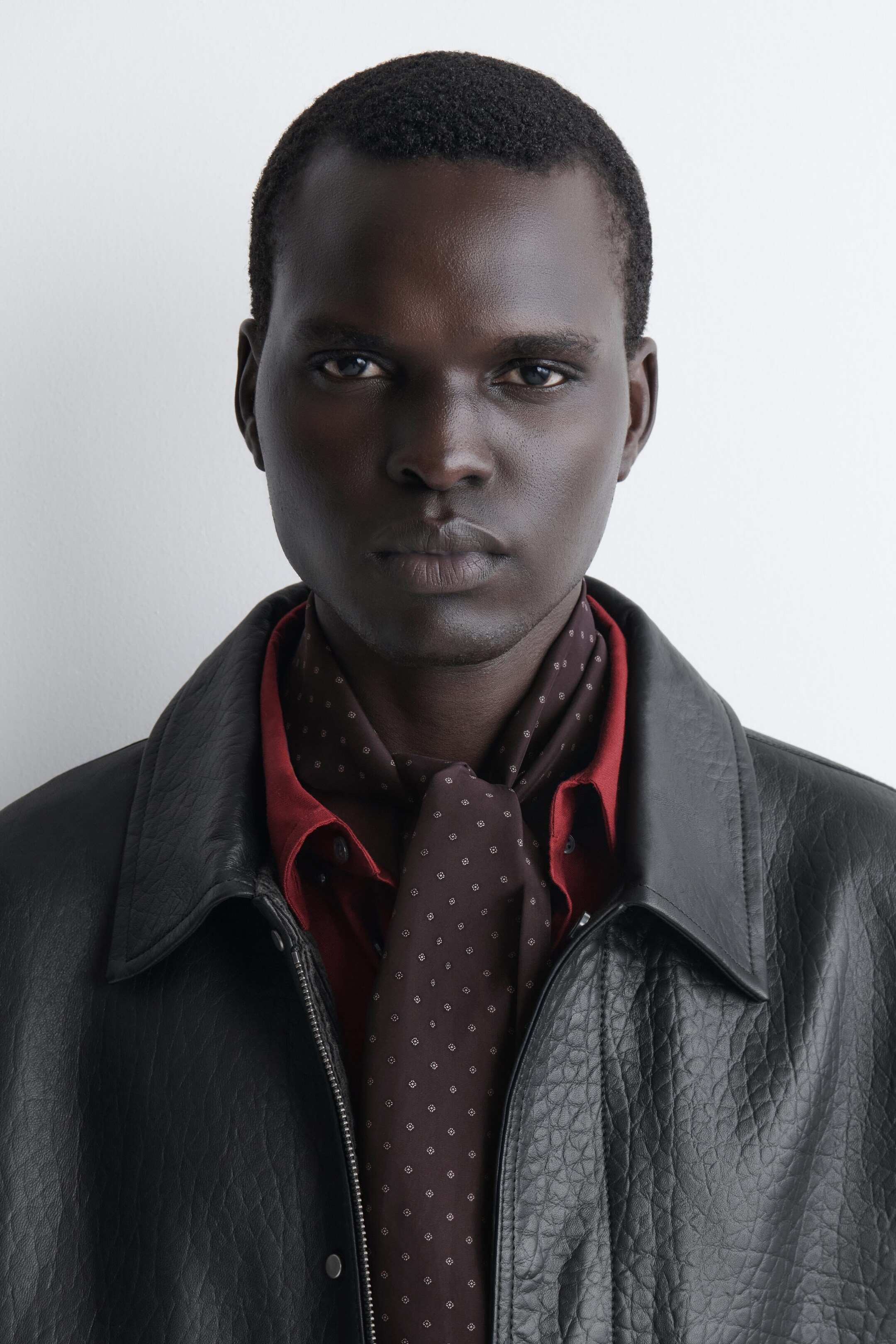 A slim, dark brown silk scarf featuring a white micro-diamond motif is shown from the front, tied around the neck and draping down the chest. It is being styled layered over a dark red collared shirt and under a textured black leather jacket. A slim, dark brown silk scarf featuring a white micro-diamond motif is shown from the front, tied around the neck and draping down the chest. It is being styled layered over a dark red collared shirt and under a textured black leather jacket.