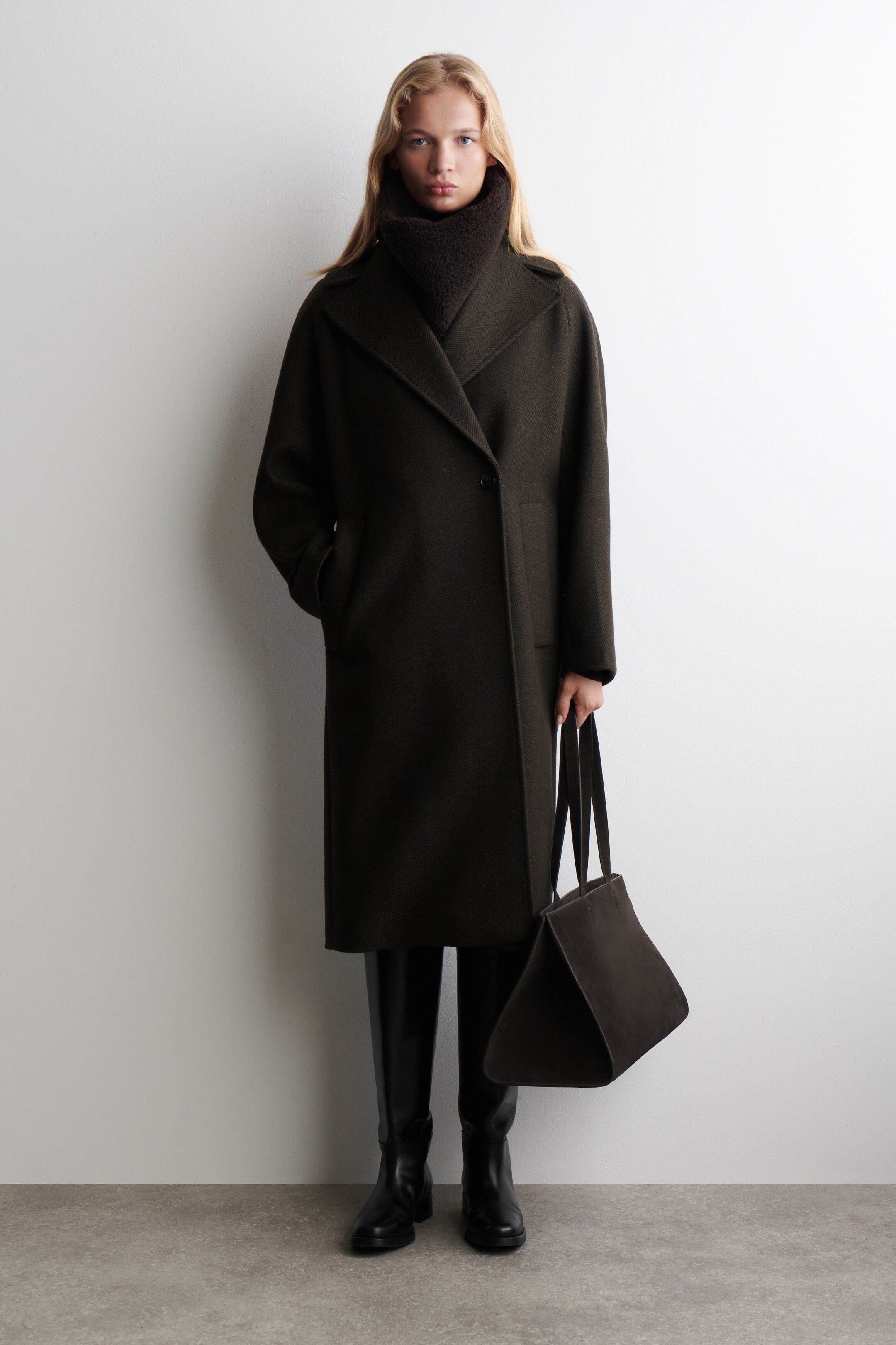 View larger image: A dark olive green long coat is featuring a relaxed fit, wide notch lapels, and a single-button closure. A dark brown textured high-neck is layering underneath the coat's collar, and jetted pockets are visible on the front.