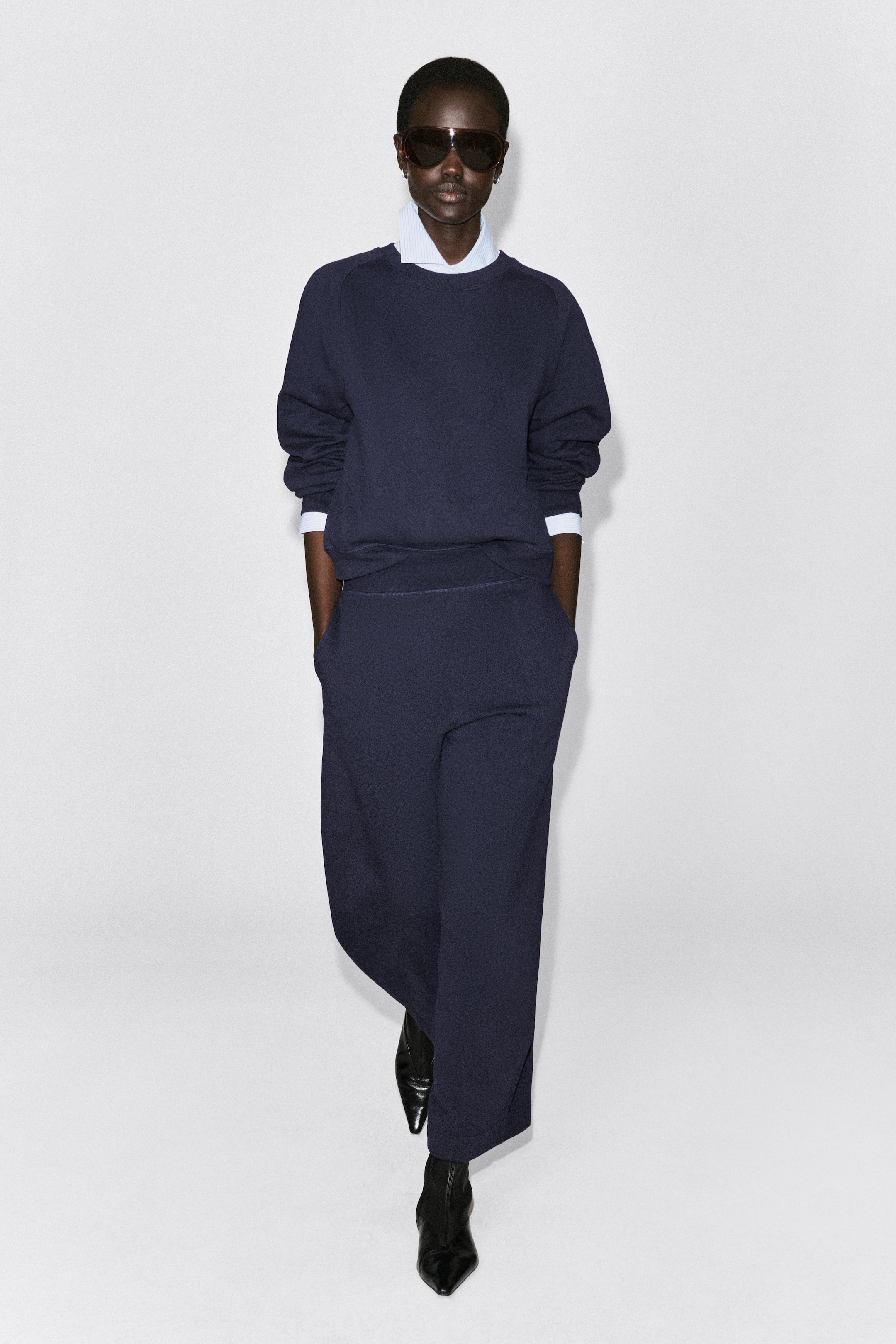 Agrandir l'image: Un jogger en coton à jambes barrel, de couleur bleu marine foncé, est présenté de face avec une silhouette décontractée et incurvée. Il présente une large ceinture et est associé à un sweat assorti ainsi qu'à des bottines noires à bout pointu.
