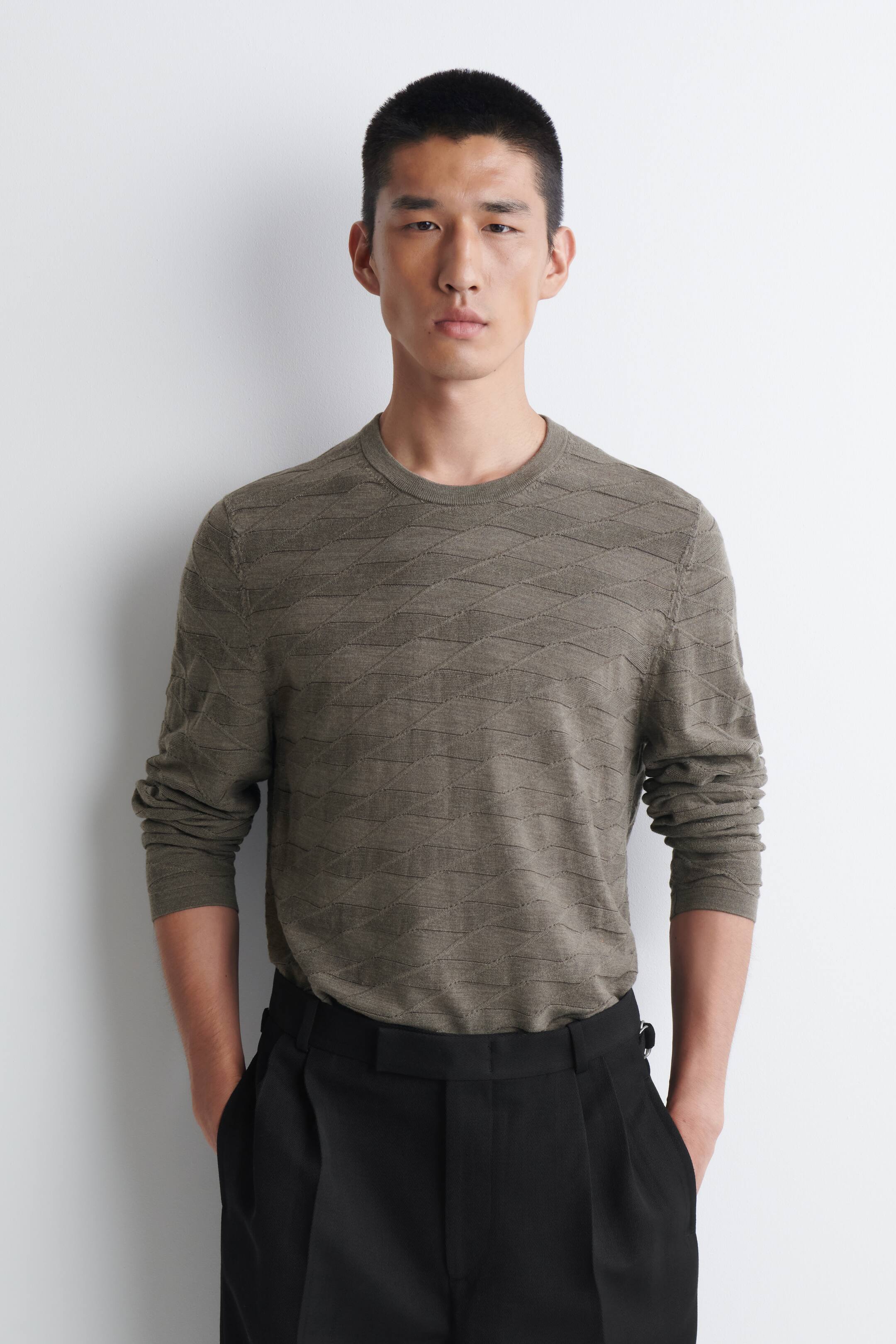 Agrandir l'image: Un pull homme vert olive est présenté de face, avec un motif tricot géométrique sur l’ensemble du corps et des manches longues, un col rond côtelé et des poignets côtelés. Sa coupe est standard, car il est rentré dans un pantalon noir.