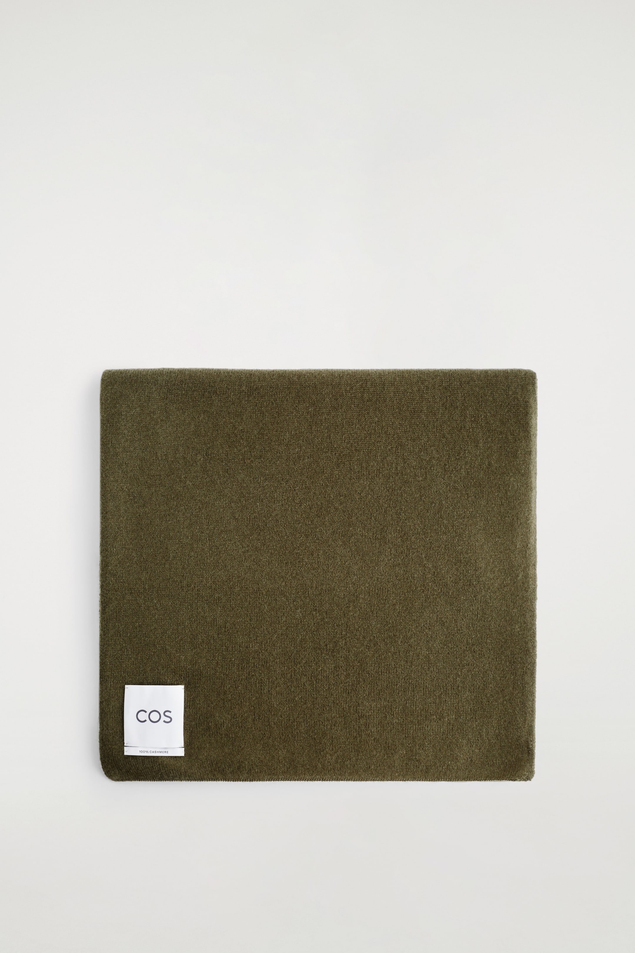 Größeres Bild anzeigen: Ein olivgrüner Kaschmirschal wird aus der Vogelperspektive präsentiert, ordentlich zu einer rechteckigen Form gefaltet. Ein weißes Etikett mit der Aufschrift "COS" und "100% CASHMERE" ist an seiner unteren linken Ecke angebracht.