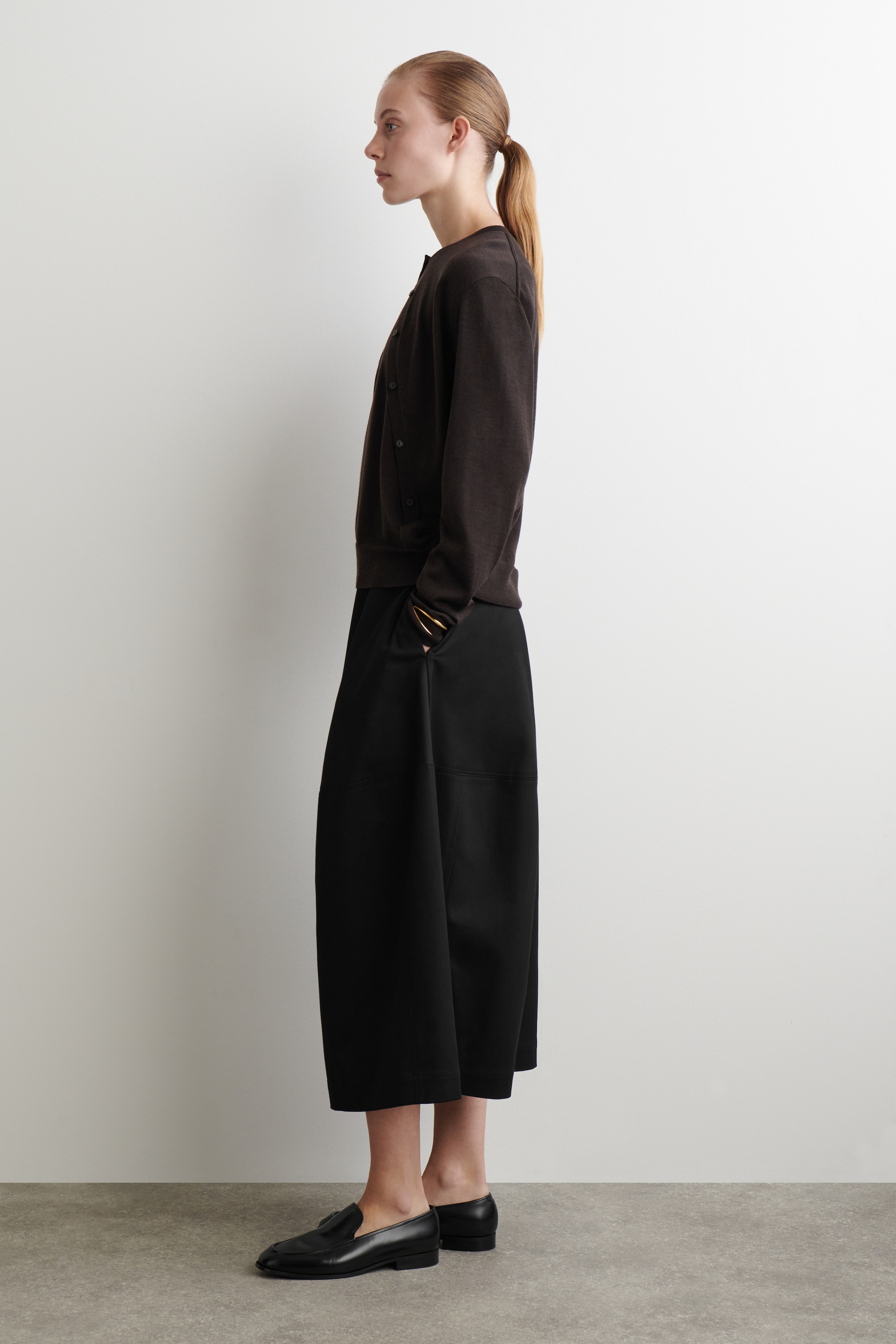 Ingrandisci l'immagine: Un cardigan marrone scuro con scollo a girocollo e bottoni sul davanti viene presentato da un'angolazione laterale, caratterizzato da una vestibilità leggermente blusata e che termina all'anca. Pantaloni neri a gamba larga e taglio corto vengono mostrati con una silhouette fluida e rilassata e cuciture frontali visibili, mentre le mani sono nelle tasche. Mocassini neri vengono indossati, mostrando un profilo classico con un tacco basso a blocco e dettagli sottili sul vamp.