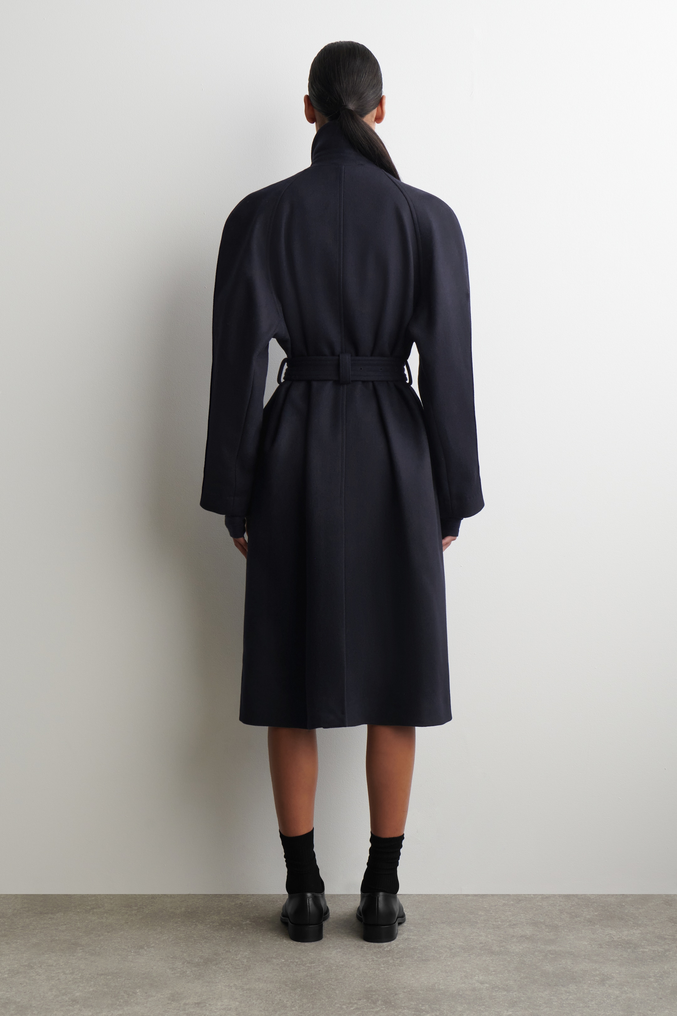 Agrandir l'image: Un manteau bleu marine foncé est présenté de dos, avec un col montant et de larges manches raglan. Le manteau est resserré à la taille avec une ceinture en tissu assorti, et son ourlet tombe sous le genou, créant une silhouette évasée.