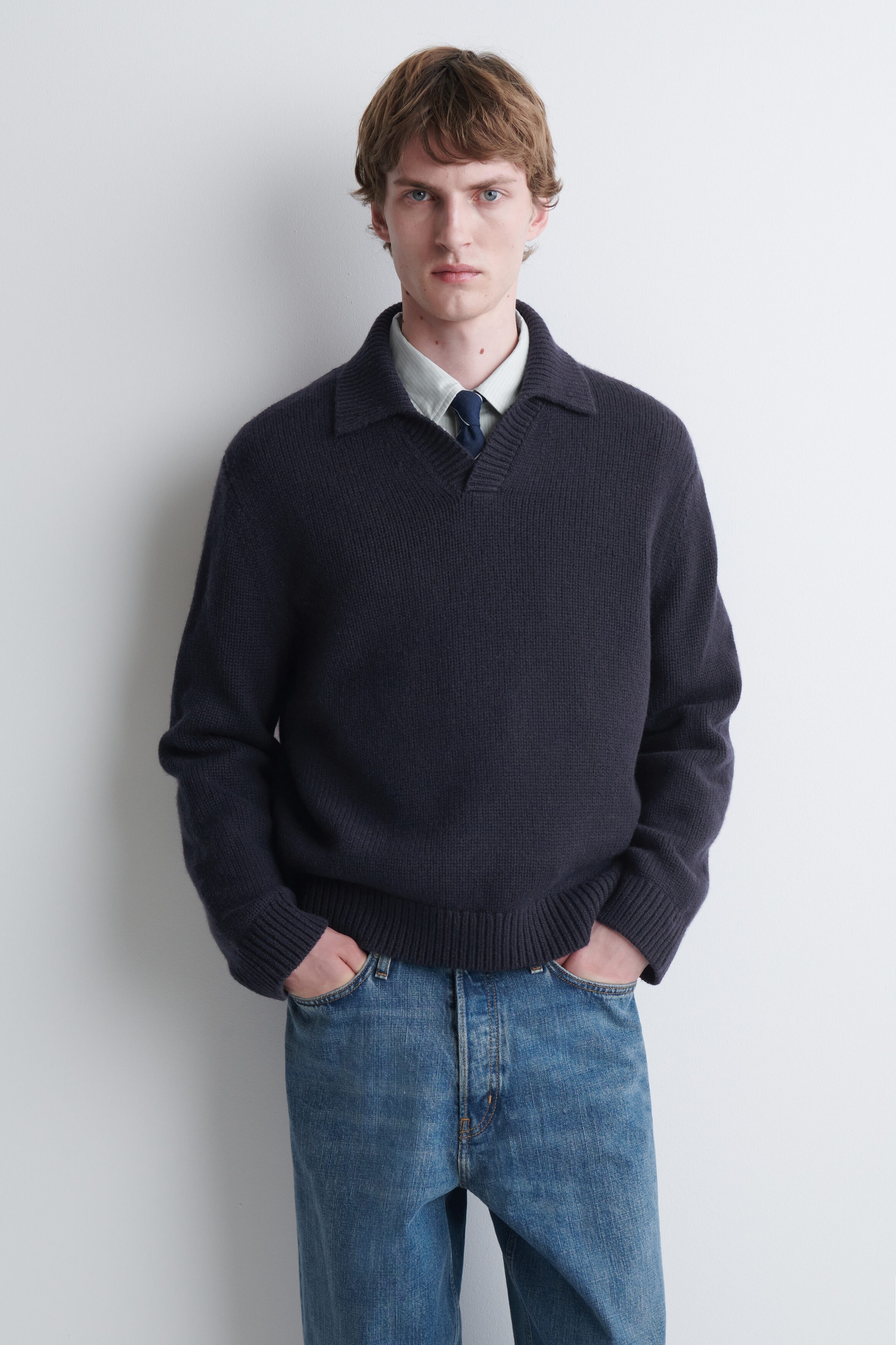 The model is wearing a dark navy blue knitted polo jumper, featuring an open collar, ribbed cuffs, and a ribbed hem. The jumper is layered over a light-coloured button-up shirt with a dark tie and paired with blue jeans. The model is wearing a dark navy blue knitted polo jumper, featuring an open collar, ribbed cuffs, and a ribbed hem. The jumper is layered over a light-coloured button-up shirt with a dark tie and paired with blue jeans.