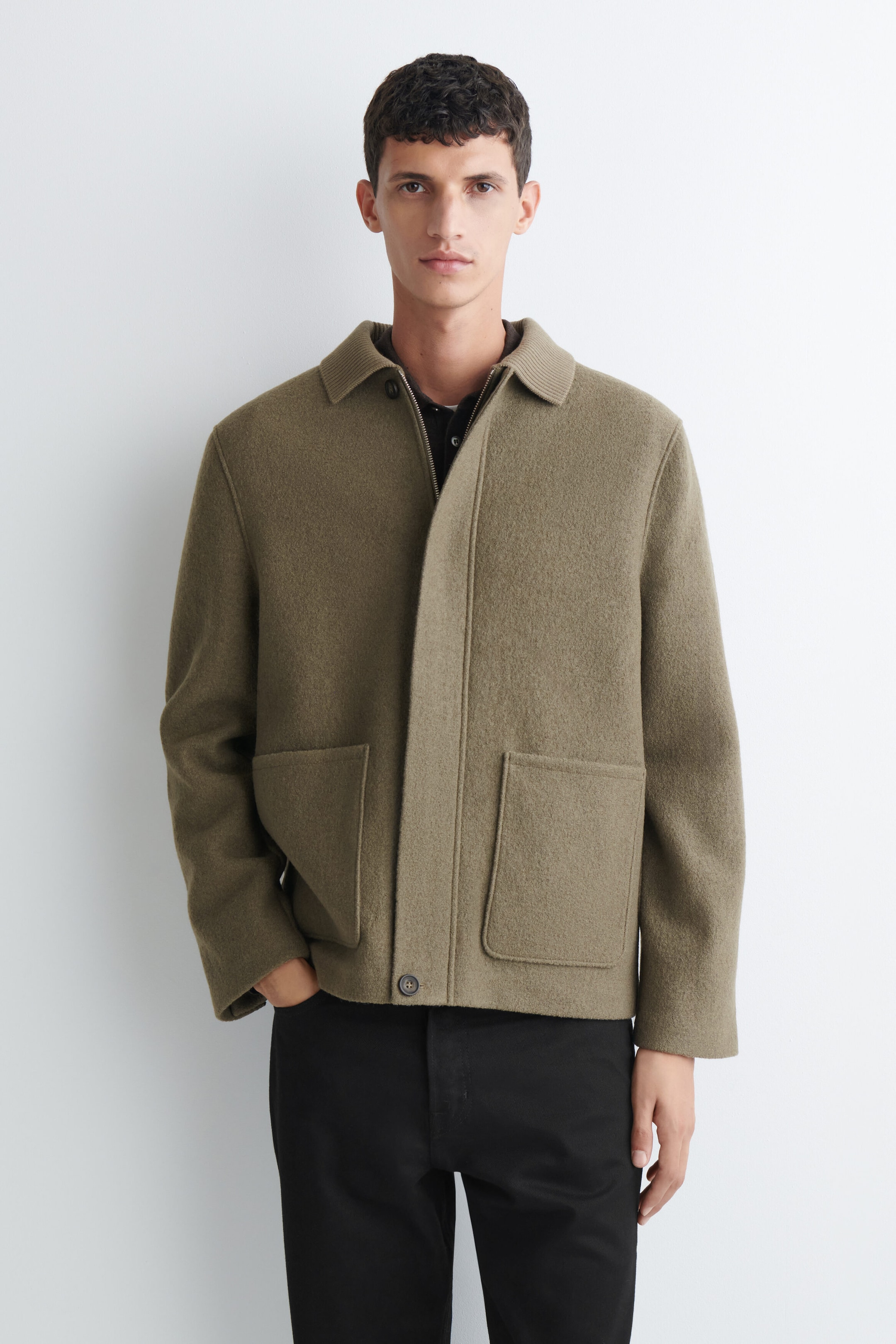 A boiled wool chore jacket in olive green, featuring a ribbed collar, concealed front closure, and two large patch pockets, is worn by the model. The jacket is layered over a dark collared shirt and paired with black trousers, with the model standing with one hand resting in a pocket. A boiled wool chore jacket in olive green, featuring a ribbed collar, concealed front closure, and two large patch pockets, is worn by the model. The jacket is layered over a dark collared shirt and paired with black trousers, with the model standing with one hand resting in a pocket.