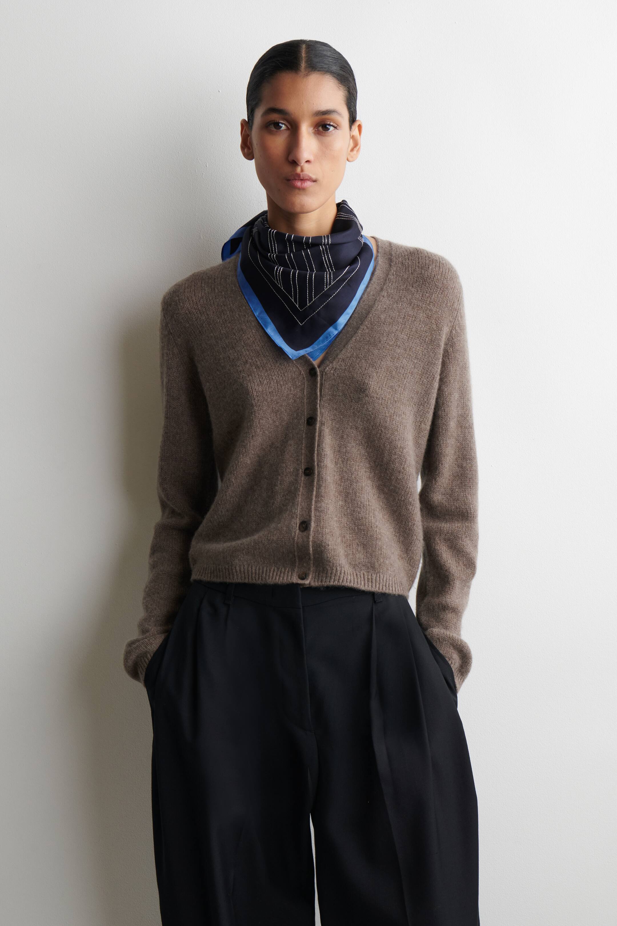 Agrandir l'image: Un cardigan en maille duveteux brun clair, légèrement raccourci, avec un col en V et des boutons foncés sur le devant, est porté sur un foulard carré bleu marine noué autour du cou et présentant un motif géométrique blanc et bleu. Un pantalon noir plissé à jambes larges est porté.