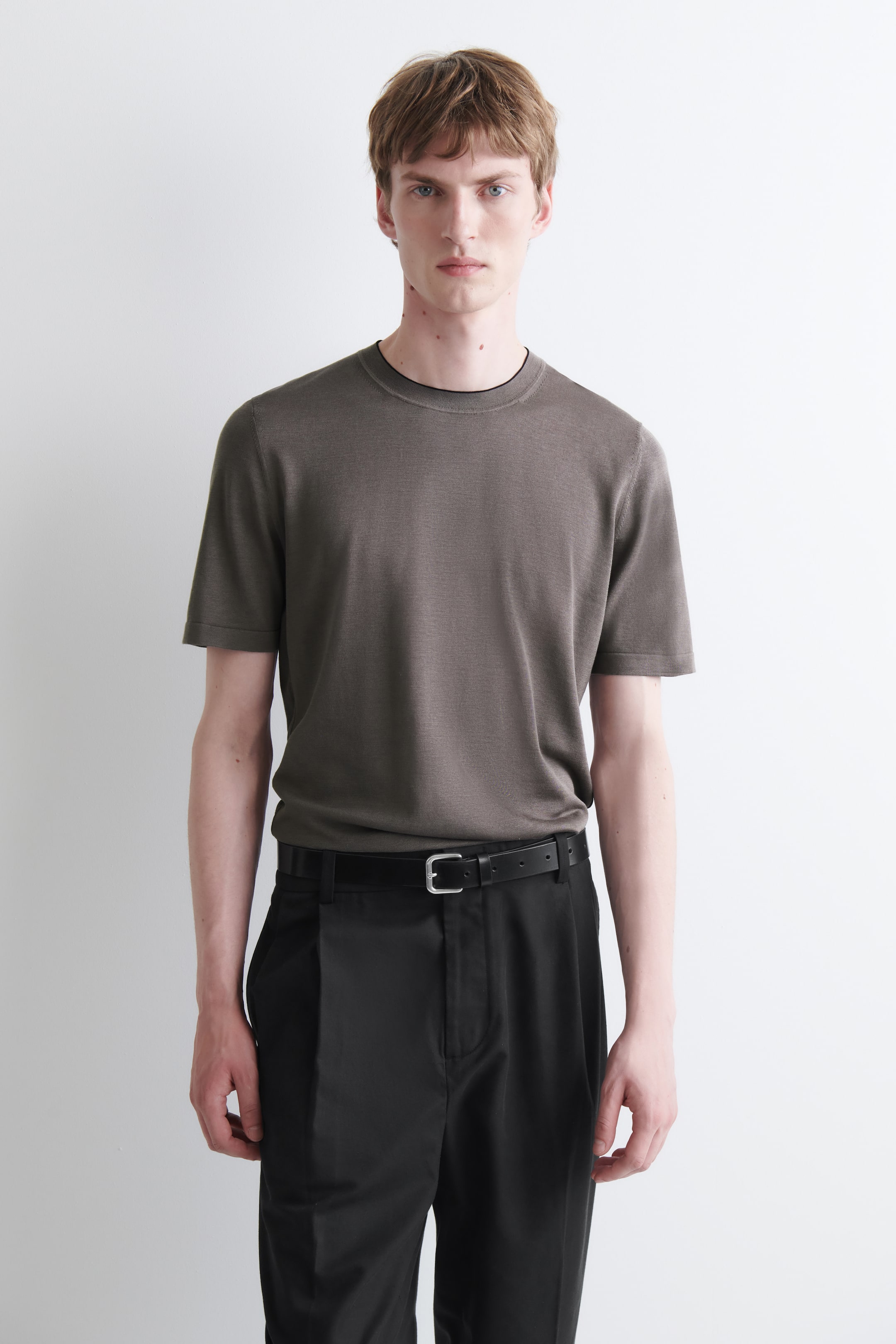 Agrandir l'image: Un T-shirt gris foncé à manches courtes présente une coupe ajustée et un liseré noir discret au niveau de son col rond. Le mannequin porte le T-shirt, rentré dans un pantalon noir.
