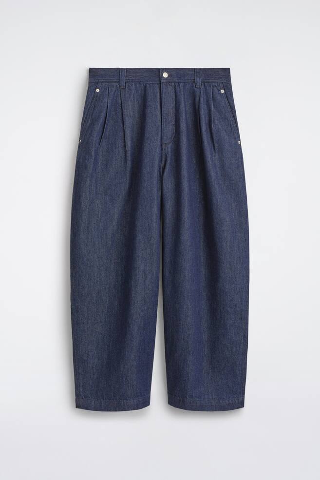 These pleated denim relaxed-fit barrel-leg trousers are shown from the front, showcasing a dark blue wash, a high-rise waistband with belt loops, and a central button fastening. The design features prominent front pleats, side slip pockets with metal rivets, and curved, relaxed-fit legs that taper slightly at the hem. - 1