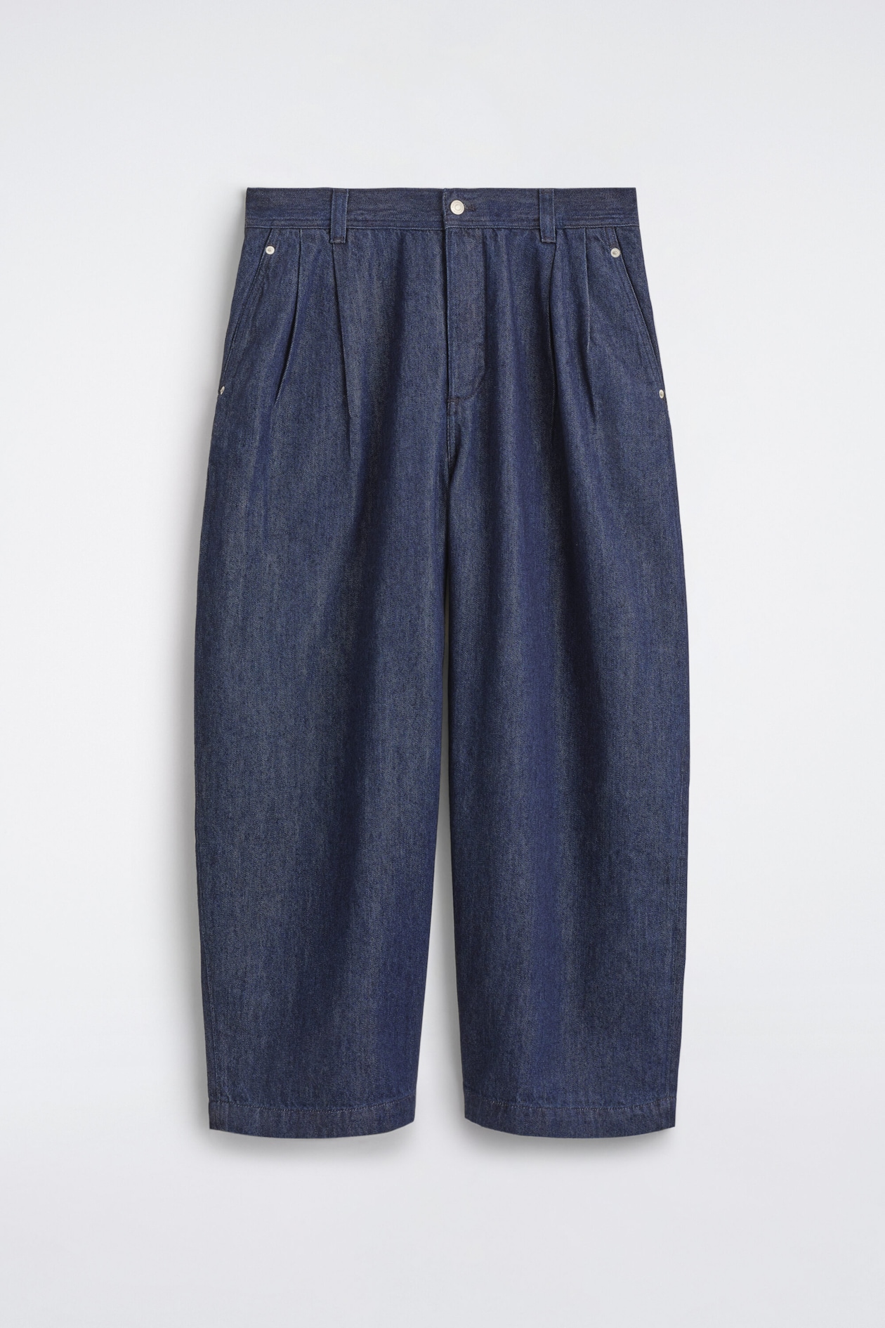 Ce pantalon barrel à jambe ample en denim plissé est présenté de face, mettant en valeur un délavage bleu foncé, une taille haute avec passants de ceinture et une fermeture centrale par bouton. Le modèle présente de marquées pinces sur le devant, des poches latérales fendues avec rivets métalliques, ainsi que des jambes arrondies à la coupe décontractée, légèrement resserrées à l’ourlet.