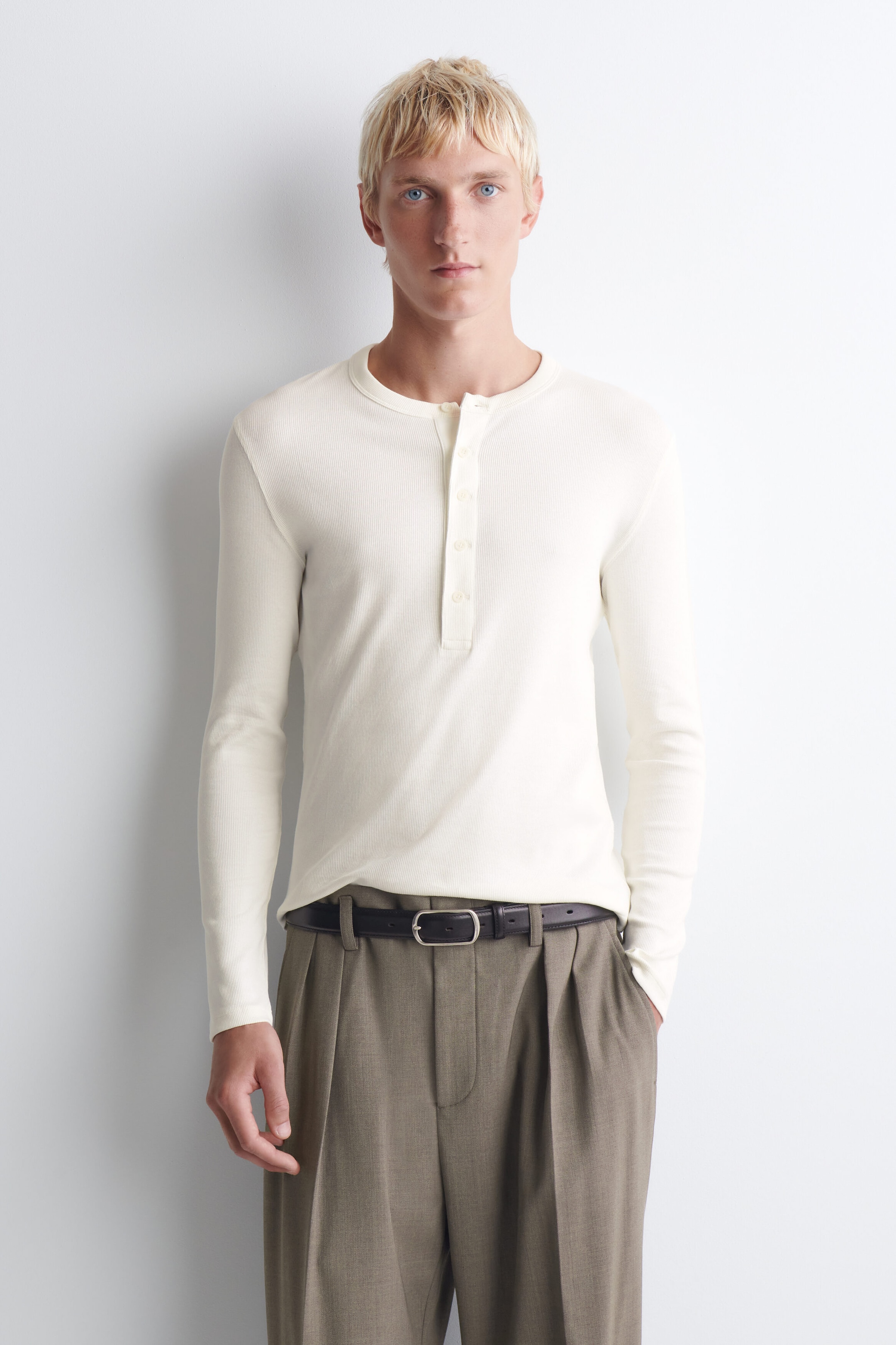 An off-white, slim-fit ribbed cotton long-sleeved Henley T-shirt is shown from the front, featuring a crew neck with a buttoned placket and a fine ribbed texture. The T-shirt is tucked into pleated trousers with a black belt, showcasing its close-fitting silhouette. An off-white, slim-fit ribbed cotton long-sleeved Henley T-shirt is shown from the front, featuring a crew neck with a buttoned placket and a fine ribbed texture. The T-shirt is tucked into pleated trousers with a black belt, showcasing its close-fitting silhouette.