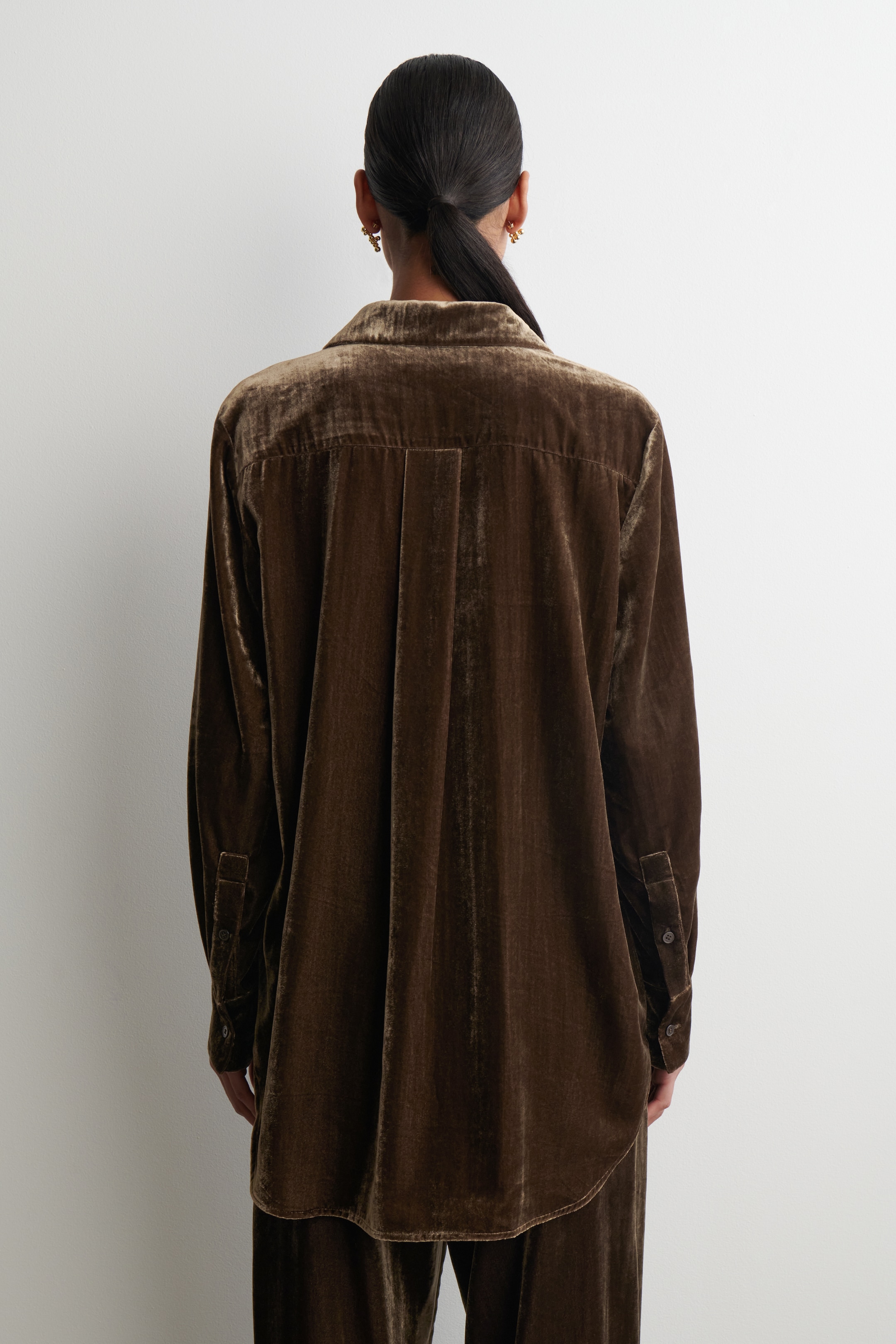 View larger image: A brown velvet shirt is being viewed from the back, featuring a classic collar, a back yoke with a central pleat, and long sleeves with buttoned cuffs. The shirt, presenting a relaxed fit, is being styled with matching velvet pants.