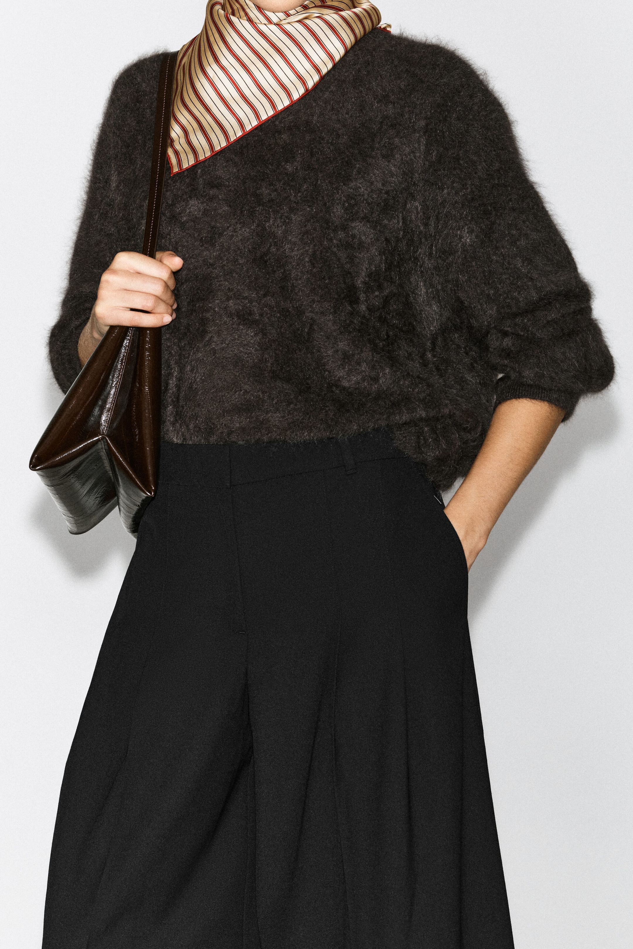 The model is wearing black pleated wool-blend wide-leg trousers, standing with one hand resting in the side pocket. The trousers feature a tailored waistband with belt loops and are styled with a textured dark grey knit jumper, a striped neck scarf, and a dark brown leather shoulder bag. The model is wearing black pleated wool-blend wide-leg trousers, standing with one hand resting in the side pocket. The trousers feature a tailored waistband with belt loops and are styled with a textured dark grey knit jumper, a striped neck scarf, and a dark brown leather shoulder bag.