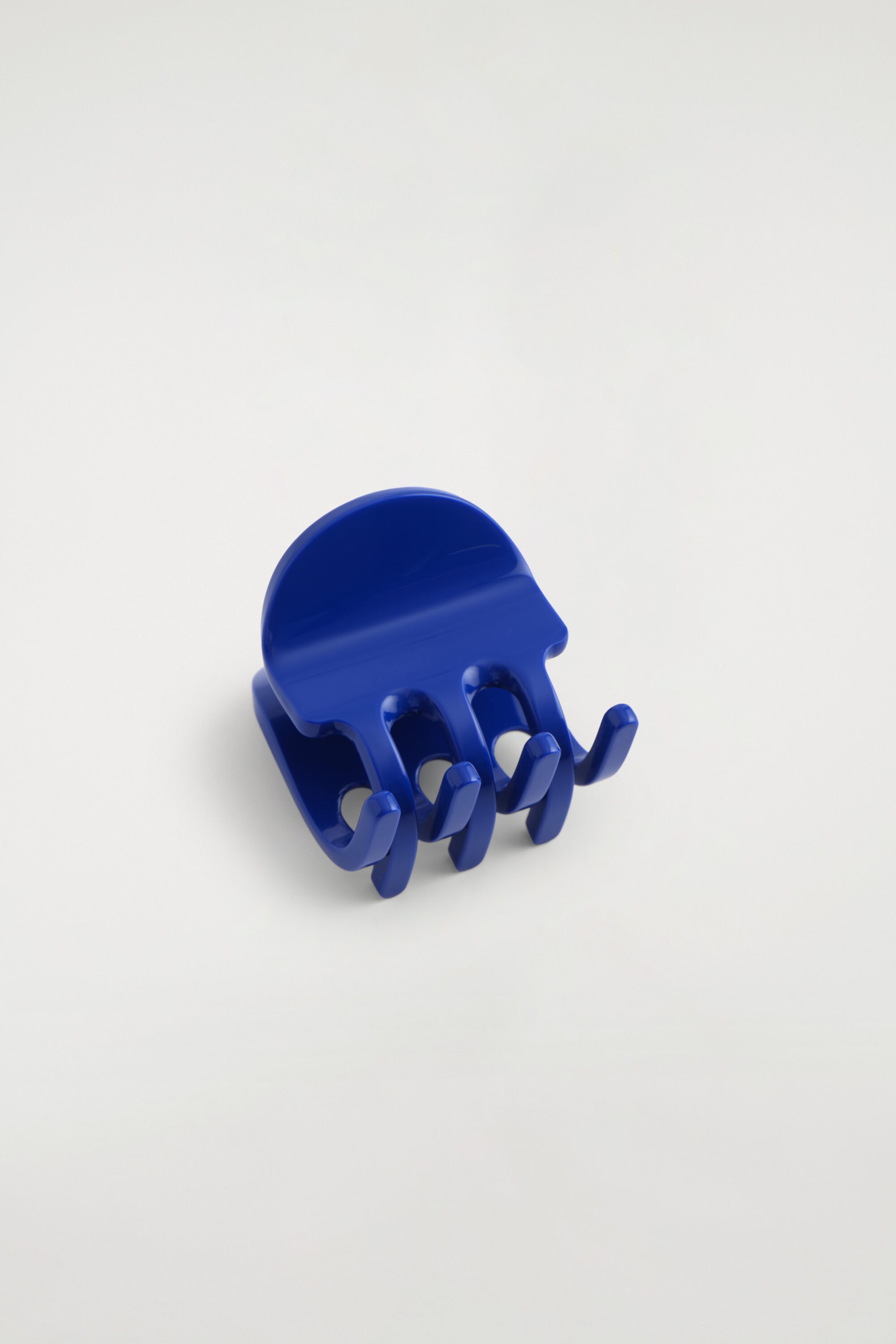 A cobalt blue hair claw clip is resting at an angle, showcasing its glossy plastic construction, semi-circular top tab, and curved interlocking teeth.