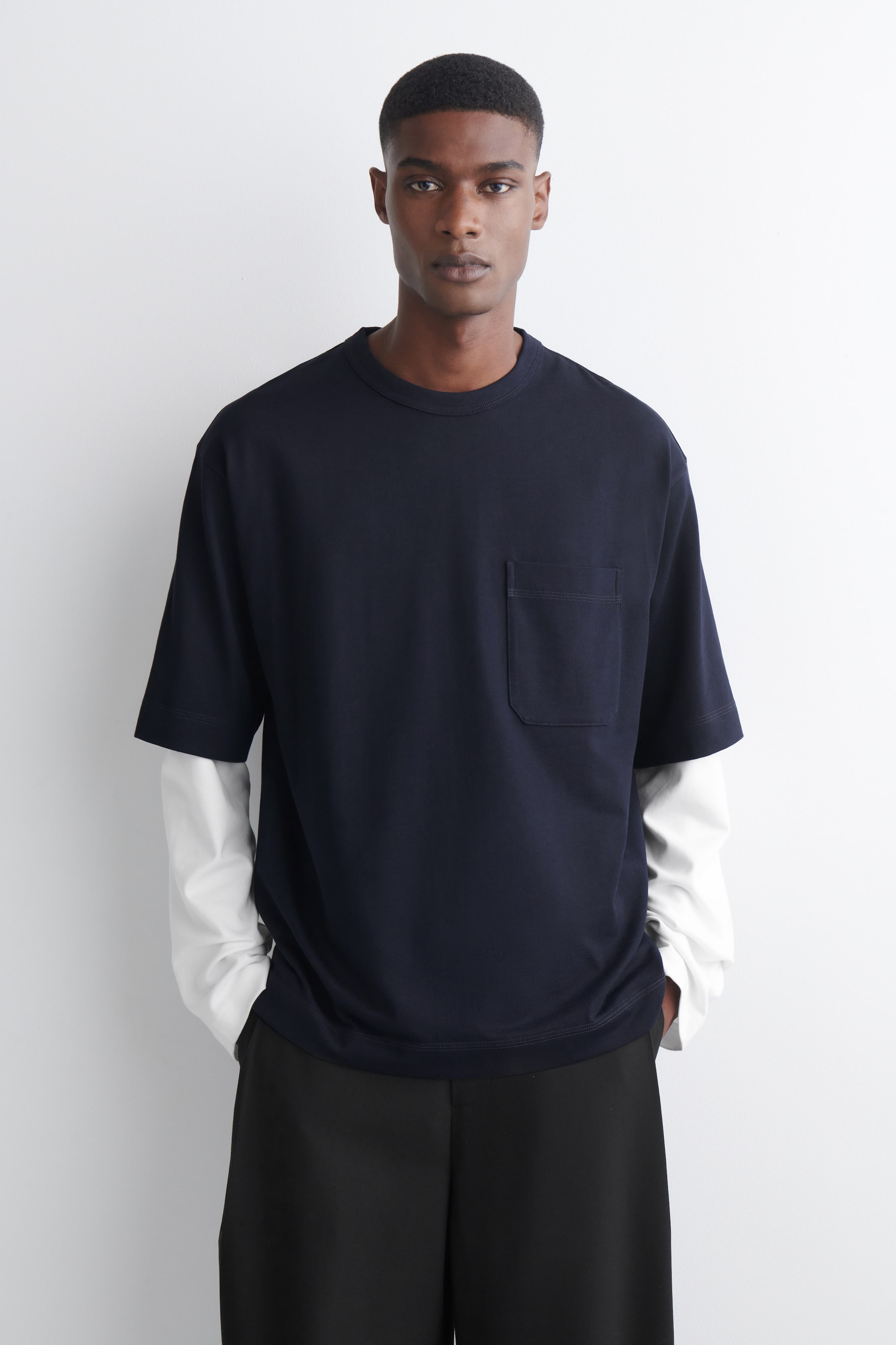 Agrandir l'image: Un t-shirt bleu foncé à manches courtes est orienté vers l'avant, avec un col rond, une poche plaquée sur la poitrine gauche et des manches larges. Il est superposé à une chemise blanche à manches longues, avec un pantalon noir visible en dessous.