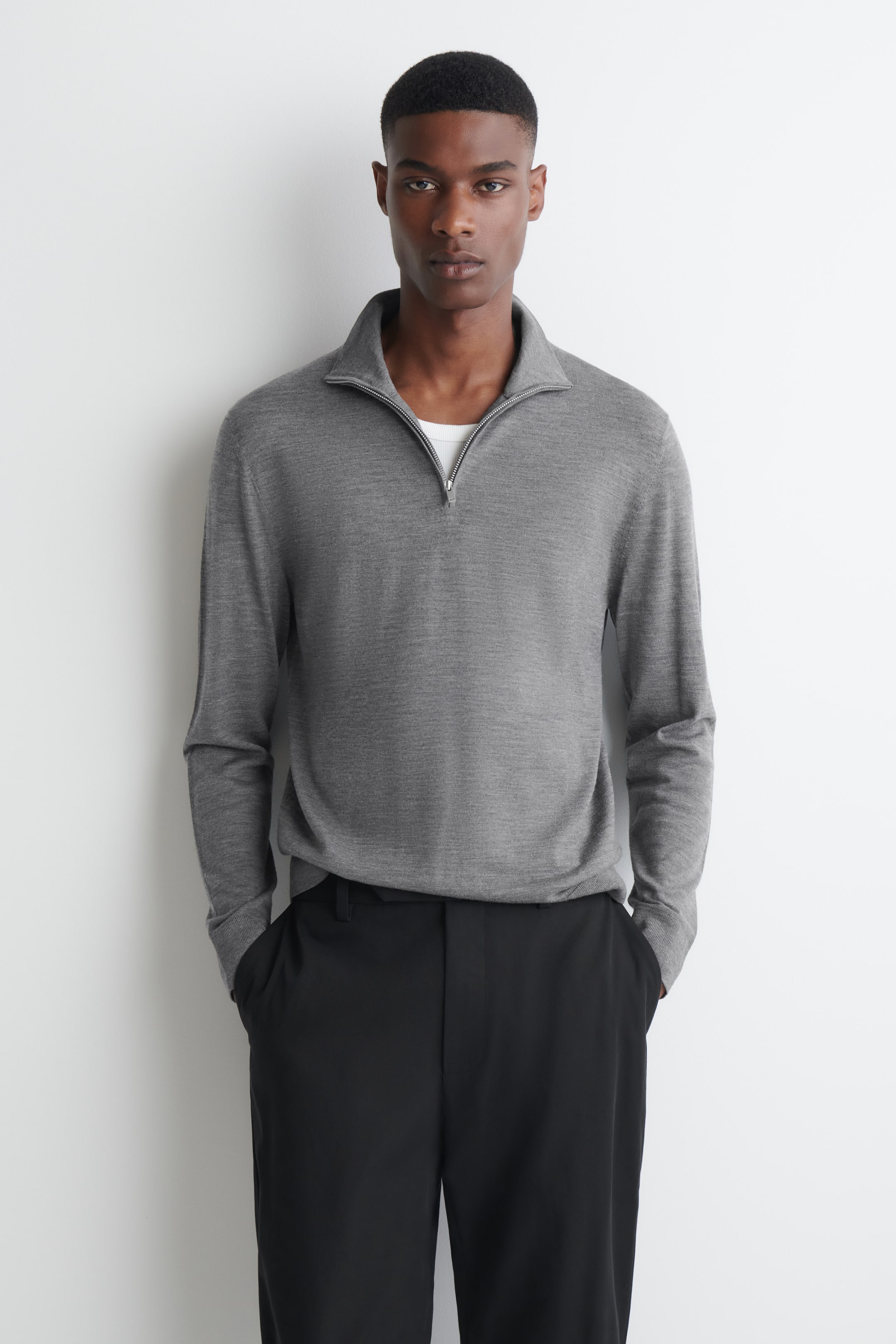 Visualizza l'immagine ingrandita: La modella indossa un maglione grigio in lana merino con collo a imbuto e zip, caratterizzato da un colletto con mezza zip argentata parzialmente aperta per mettere in risalto una t-shirt bianca a strati. Il maglione è abbinato a pantaloni neri, con la modella che tiene le mani nelle tasche.