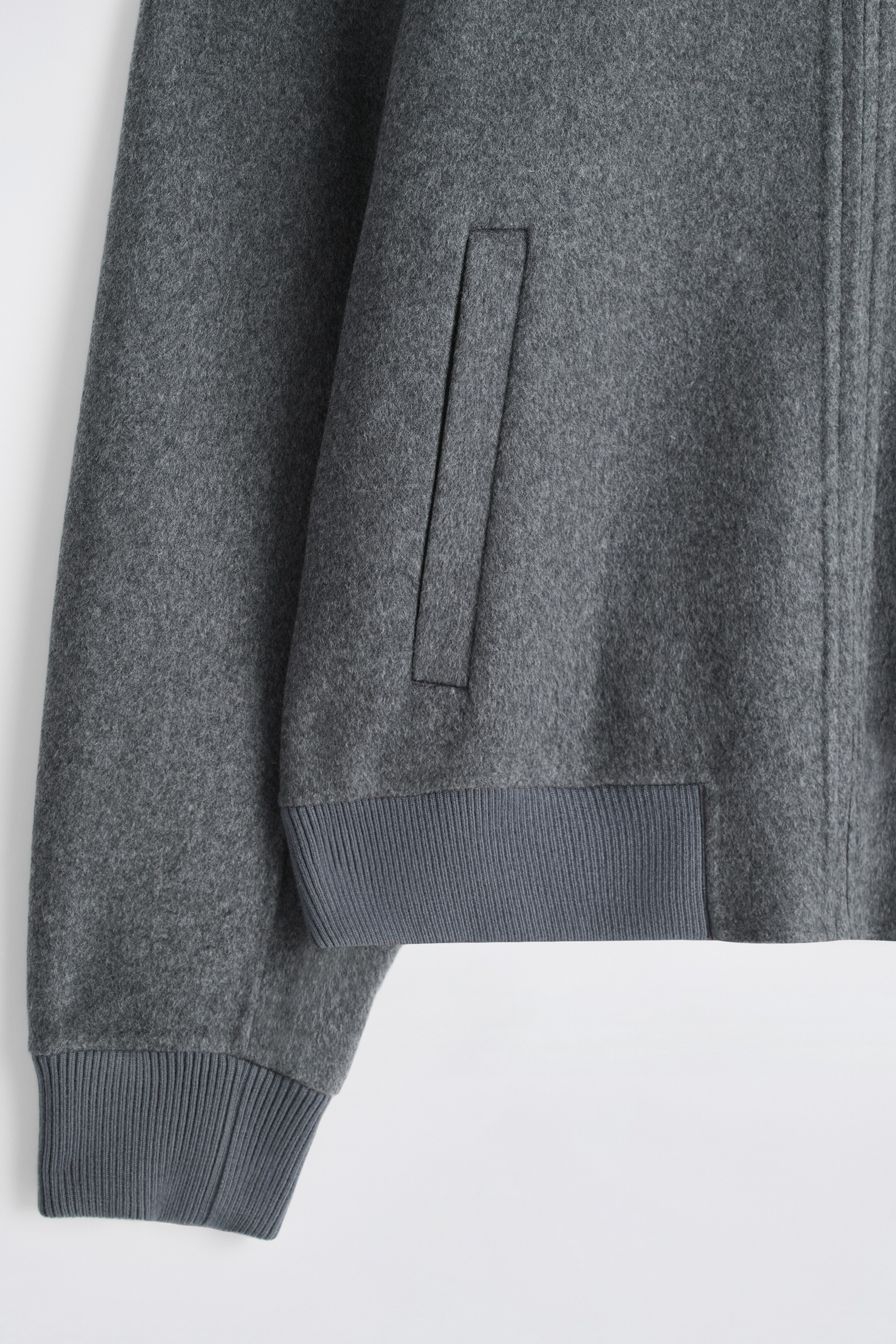 View larger image: A grey bomber jacket is presenting its front left side, featuring a textured fabric, a ribbed cuff on the long sleeve, and a matching ribbed hem. A slanted welt pocket is visible on the front, alongside a vertical seam indicating the main closure.