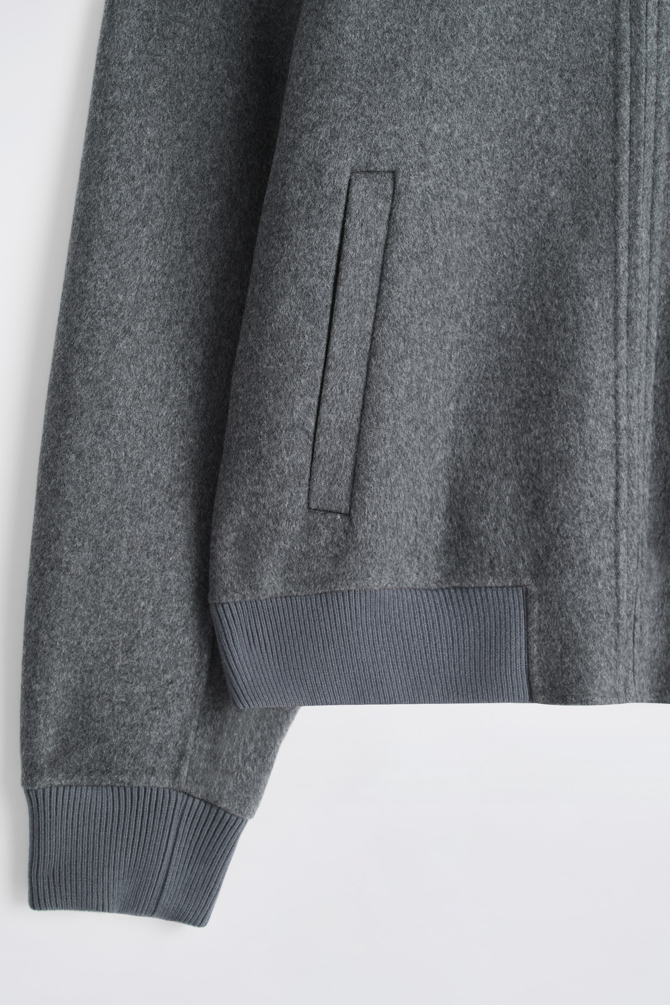 Ver imagen más grande: Una chaqueta tipo bomber gris está mostrando su lado izquierdo frontal, con una tela texturizada, un puño acanalado en la manga larga y un dobladillo acanalado a juego. Un bolsillo de ojal sesgado es visible en el frente, junto con una costura vertical que indica el cierre principal.