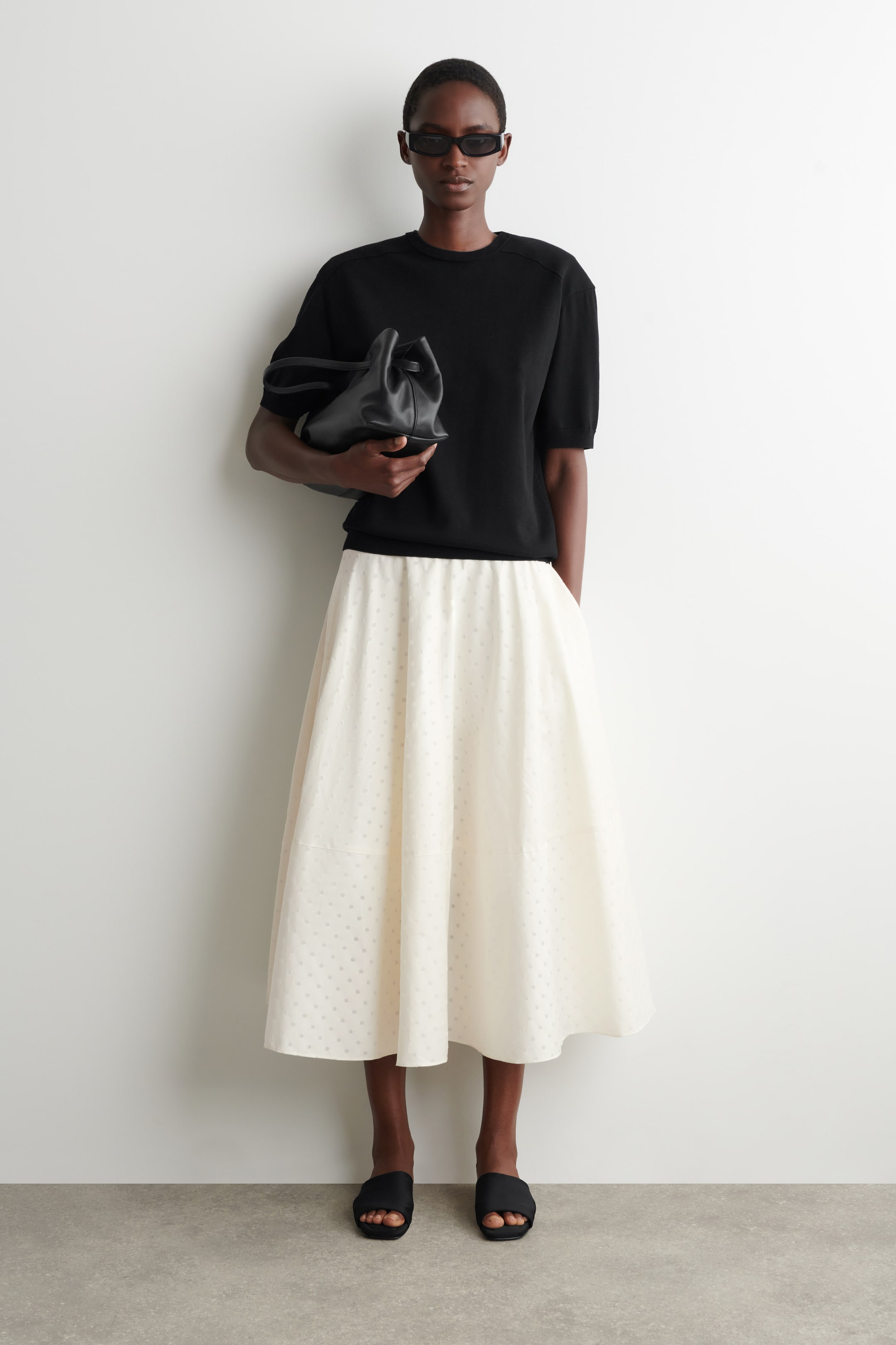 Visa större bild: En off-white midikjol med en subtil prickig jacquardväv, en utsvängd cirkelskuren silhuett och synliga toppstickade sömmar bärs av modellen. Modellen kombinerar kjolen med en svart kortärmad topp.