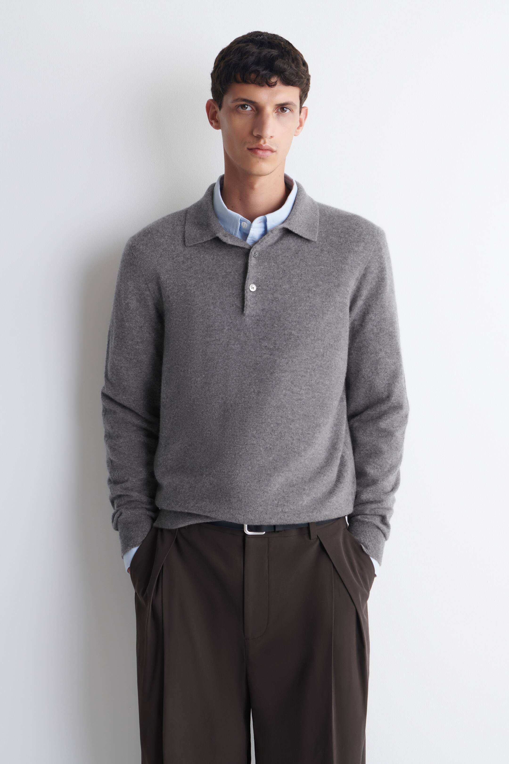Visualizza immagine più grande: Il modello indossa un maglione polo in maglia grigia sopra una camicia azzurra con colletto. Il maglione presenta un colletto e una chiusura a due bottoni con bottoni chiari, con il bottone superiore slacciato.