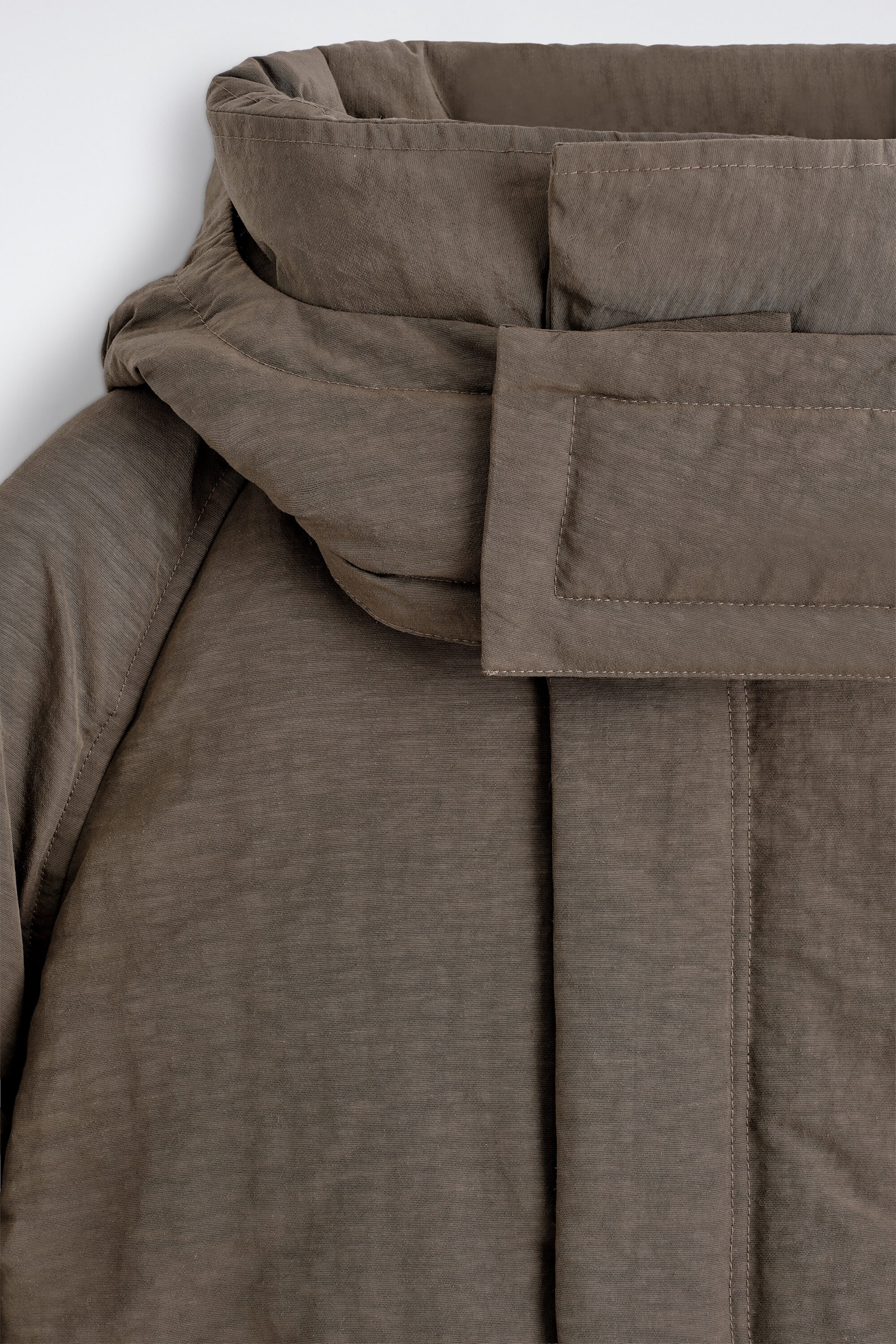 This close-up front view of a brown puffer jacket highlights the high padded collar, a wide storm flap with rectangular stitching, and a concealed front placket. The garment features a textured matte fabric with clean stitching details around the neckline and shoulders. This close-up front view of a brown puffer jacket highlights the high padded collar, a wide storm flap with rectangular stitching, and a concealed front placket. The garment features a textured matte fabric with clean stitching details around the neckline and shoulders.