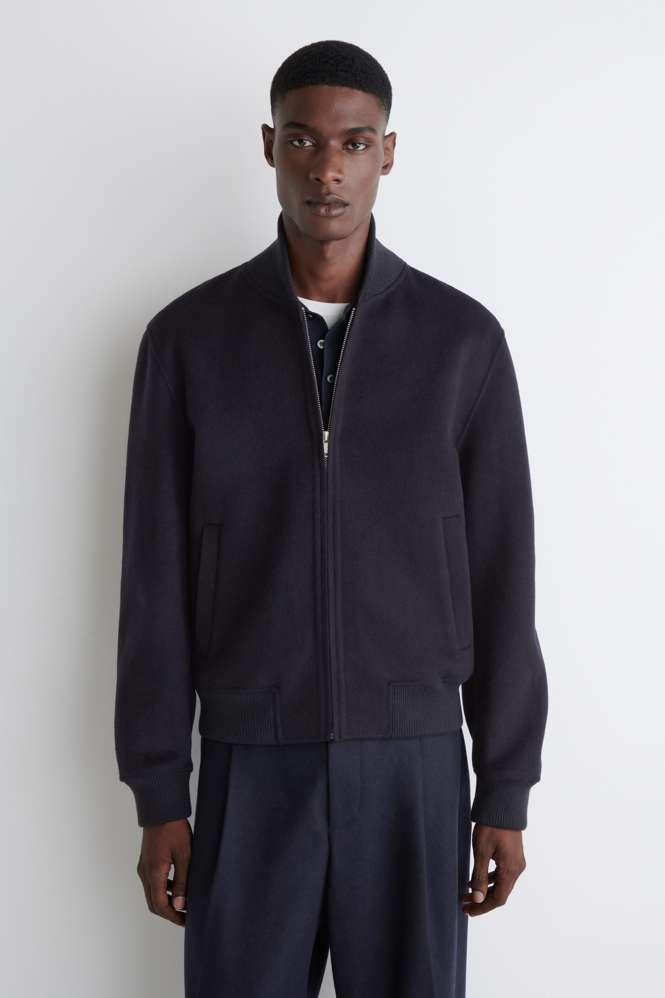 View larger image: A dark blue bomber jacket is being shown from a frontal view. It is featuring a ribbed stand-up collar, a silver-toned front zipper closure, and two front welt pockets. The jacket is completed with ribbed cuffs and a ribbed hem, and is being worn over a lighter-colored collared shirt.