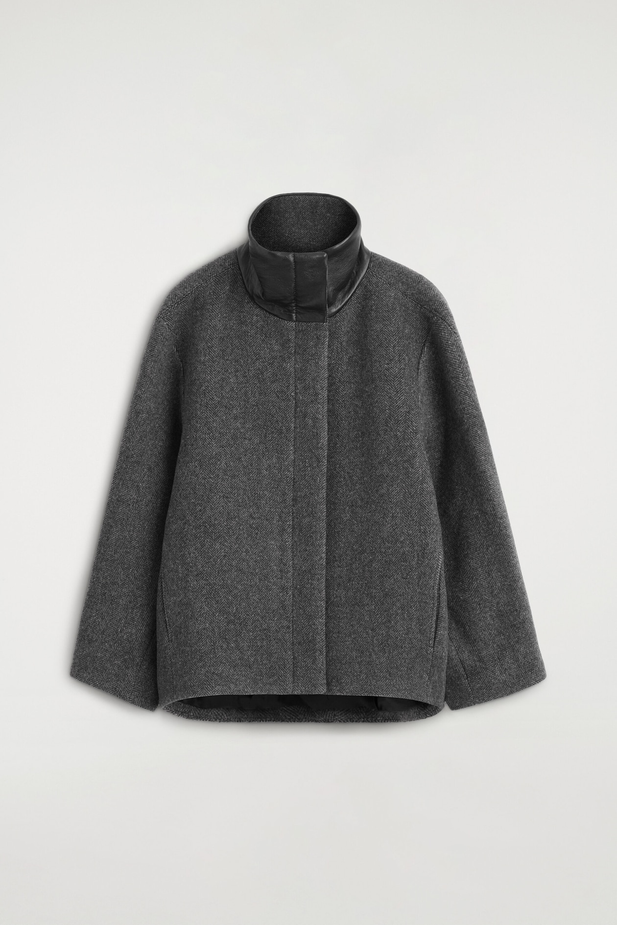 Diese graue Jacke aus Wollmischung im Fischgrätmuster ist von vorne zu sehen und zeichnet sich durch eine Oversized-Silhouette mit weiten Ärmeln aus. Sie ist mit einem hohen Stehkragen gestaltet, der durch einen schwarzen Lederkragen akzentuiert wird, sowie mit einer verdeckten Knopfleiste vorne.