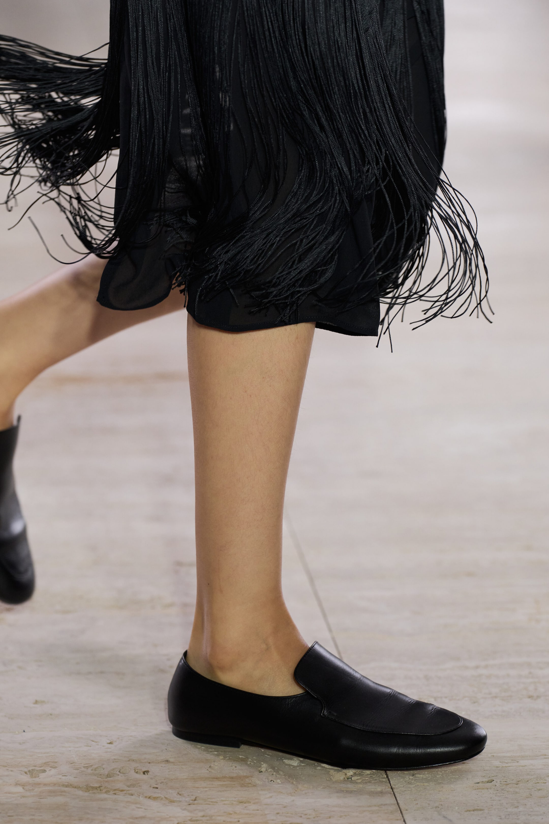 A pair of black leather loafers is shown in side profile as a model walks, featuring a streamlined almond-toe shape and a topstitched apron that sits high on the vamp. The flat shoes are paired with a black fringed skirt, showcasing their low-profile design and soft leather construction. A pair of black leather loafers is shown in side profile as a model walks, featuring a streamlined almond-toe shape and a topstitched apron that sits high on the vamp. The flat shoes are paired with a black fringed skirt, showcasing their low-profile design and soft leather construction.