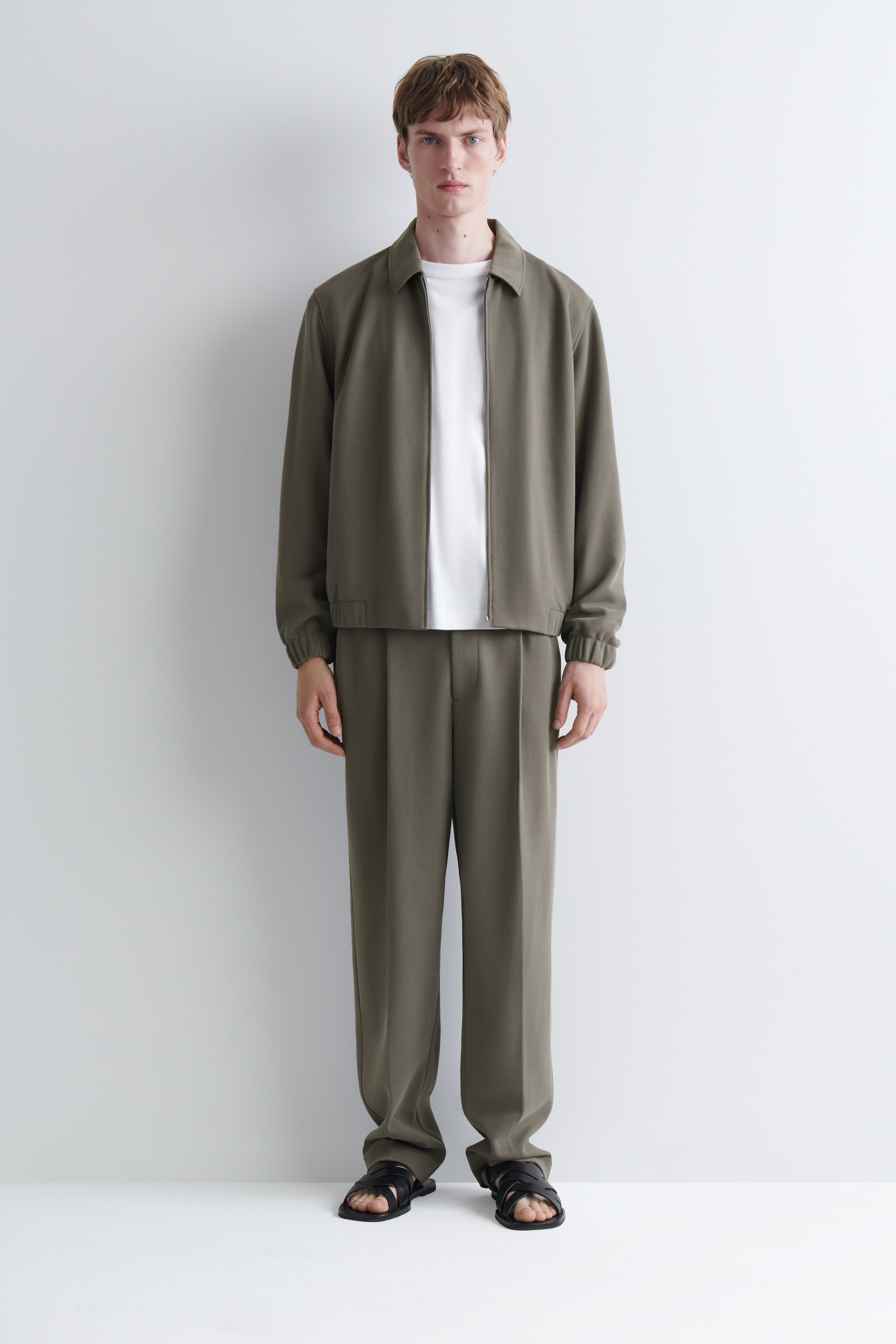 View larger image: The model is wearing an olive green collared jacket with a full zip fastening and matching pleated trousers. A white crew neck T-shirt is layered underneath the jacket, and black strappy sandals are worn.