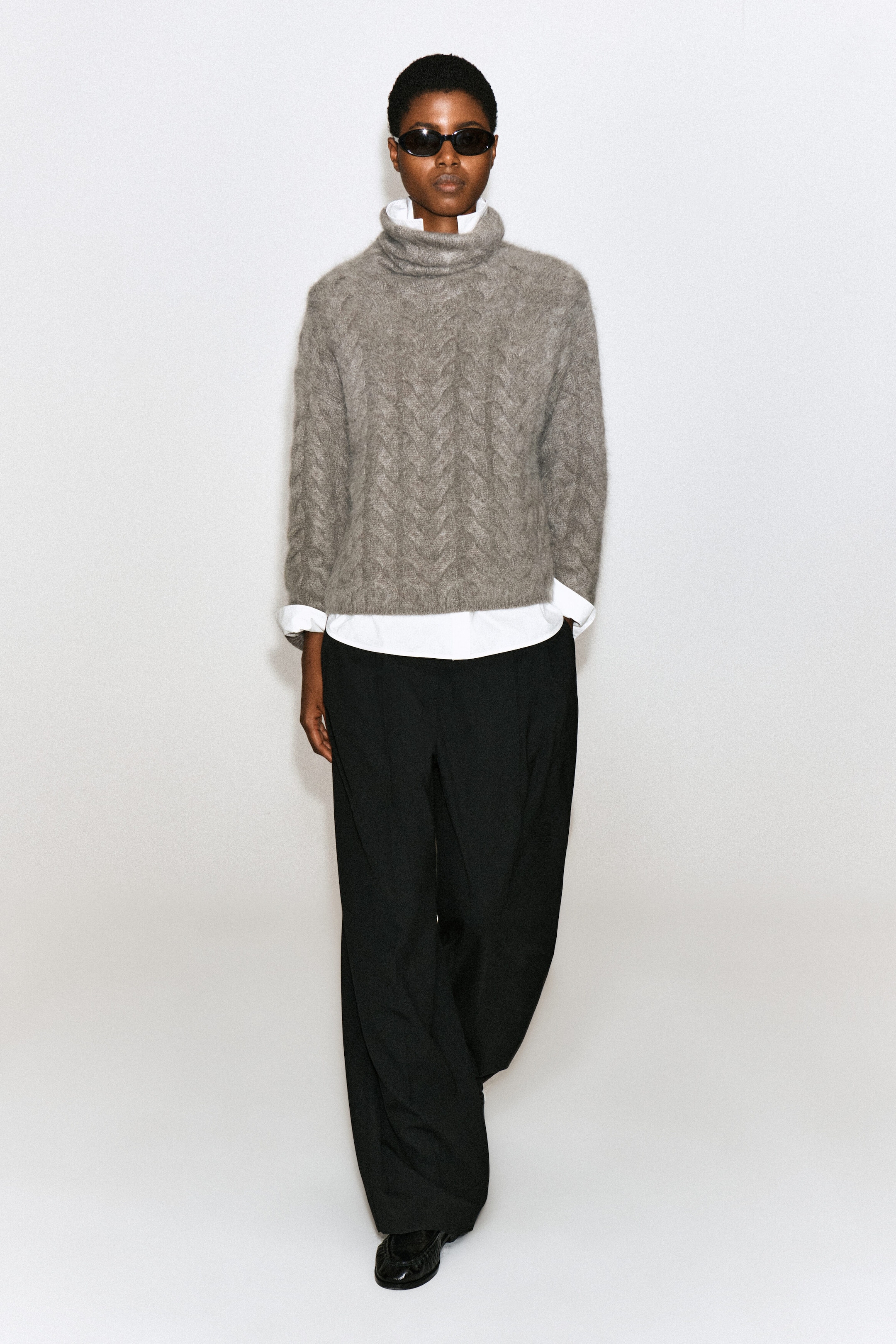 Le mannequin porte un pull gris à col montant en cachemire brossé à torsades, mettant en valeur son motif texturé et sa coupe décontractée. Le pull est porté en superposition sur une chemise blanche à col et agencé avec un pantalon noir à jambes larges. Le mannequin porte un pull gris à col montant en cachemire brossé à torsades, mettant en valeur son motif texturé et sa coupe décontractée. Le pull est porté en superposition sur une chemise blanche à col et agencé avec un pantalon noir à jambes larges.