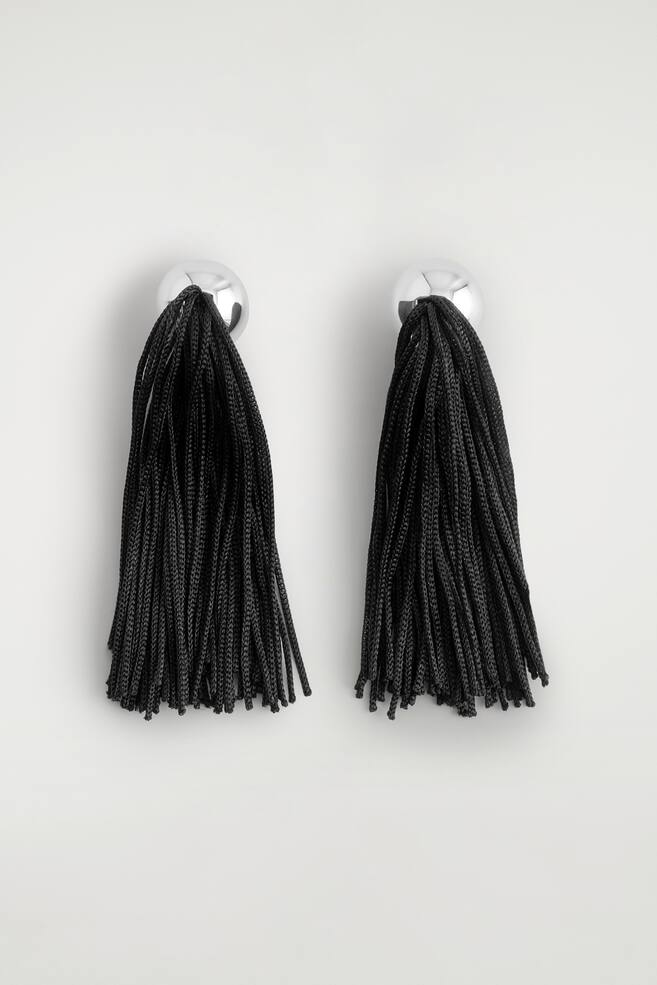 Een paar oorbellen met kwastjes wordt van voren getoond, met bovenaan gepolijste, zilverkleurige bolvormige studs. Aan elke stud hangen lange, volle kwastjes van getextureerde, zwart gevlochten koorden. - 1