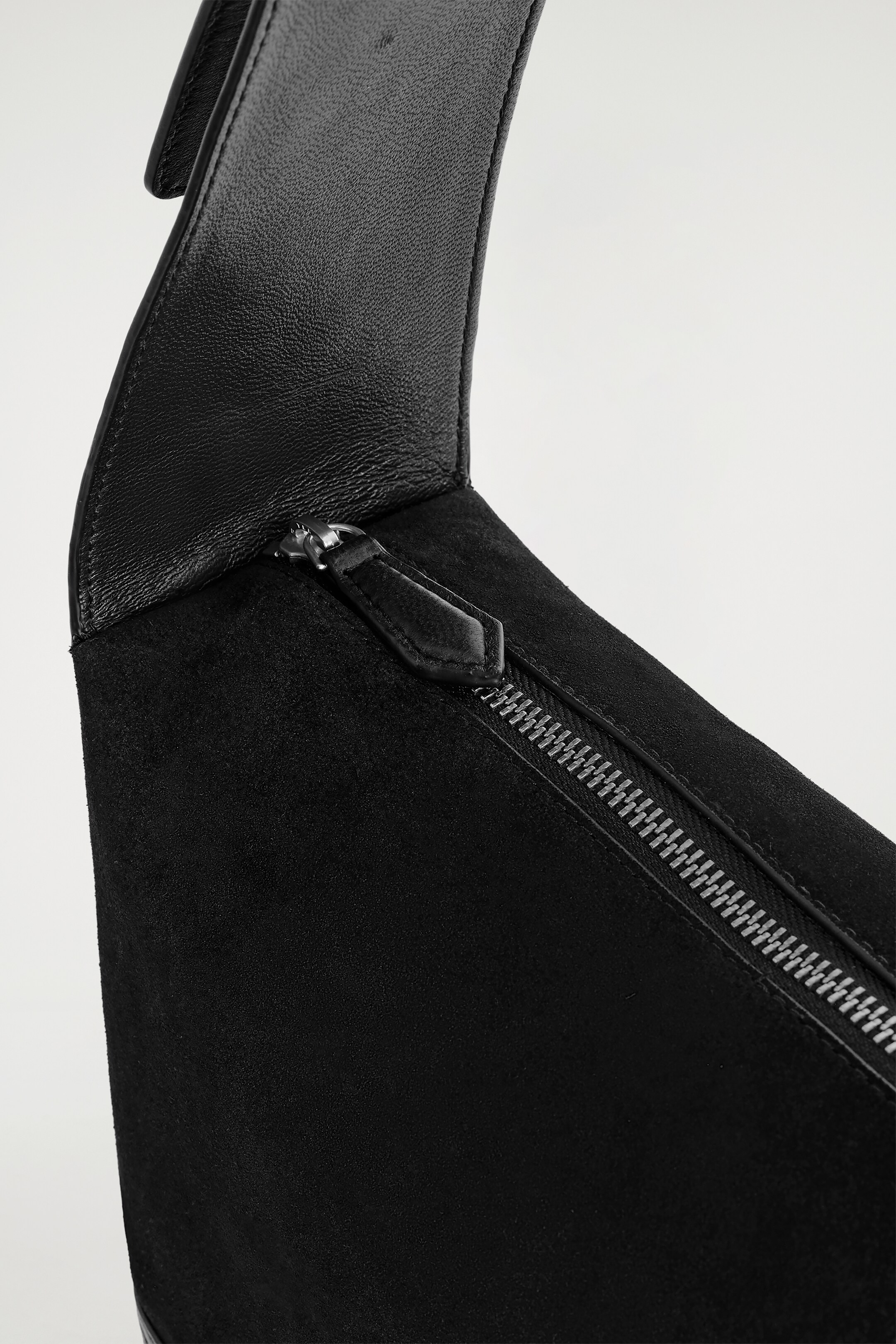 Visualizza immagine più grande: Una borsa nera è presentata da una vista dall'alto, con una sezione superiore in pelle nera liscia che si curva delicatamente e si trasforma in un corpo in camoscio nero testurizzato, dove una cerniera argentata con un tiretto in pelle nera corre in diagonale.