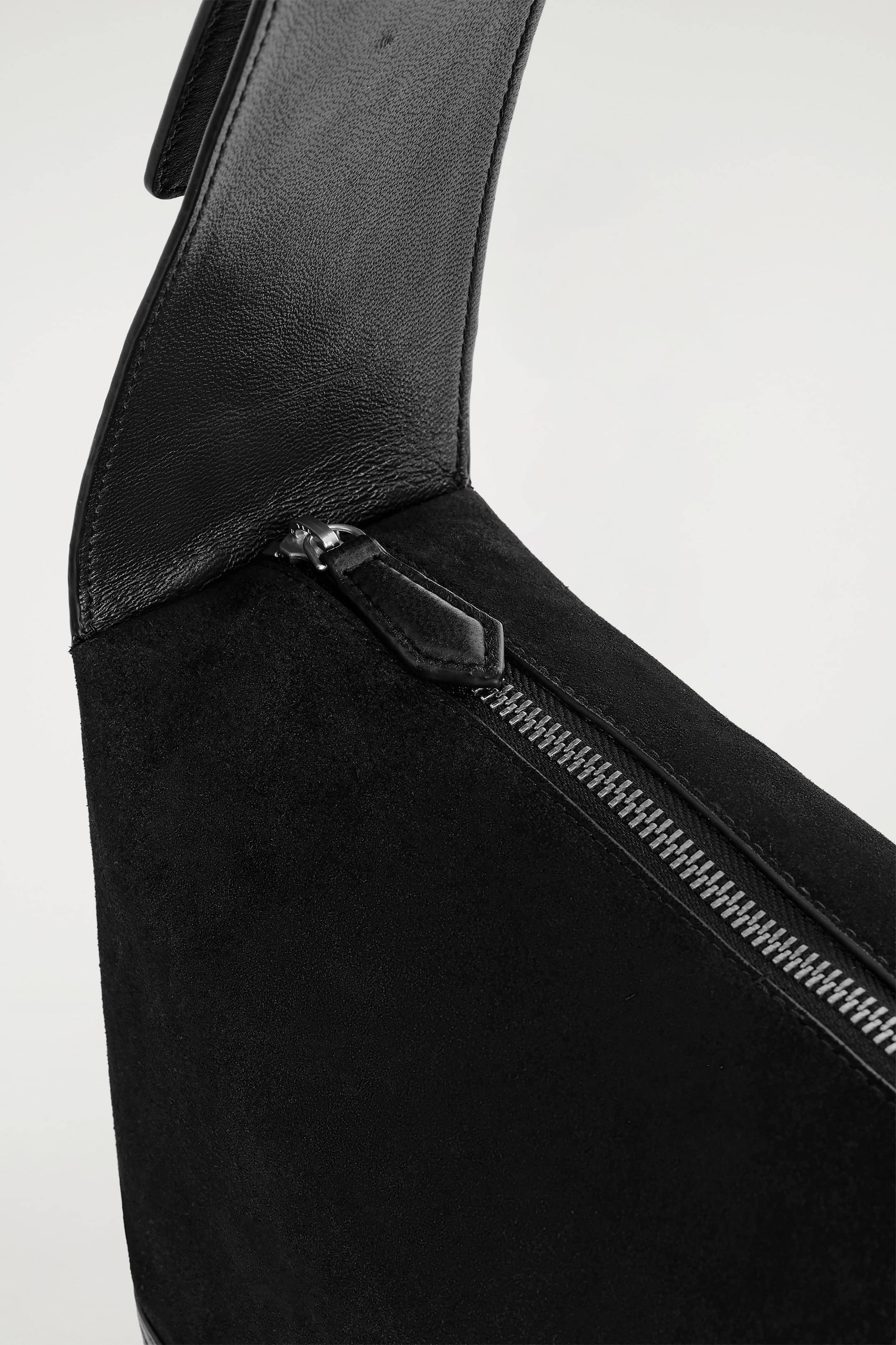 Agrandir l'image: Un sac noir est présenté en vue de dessus, avec une partie supérieure en cuir noir lisse qui se courbe doucement et se transforme en un corps en daim noir texturé, où une fermeture éclair argentée avec une languette en cuir noir court en diagonale.