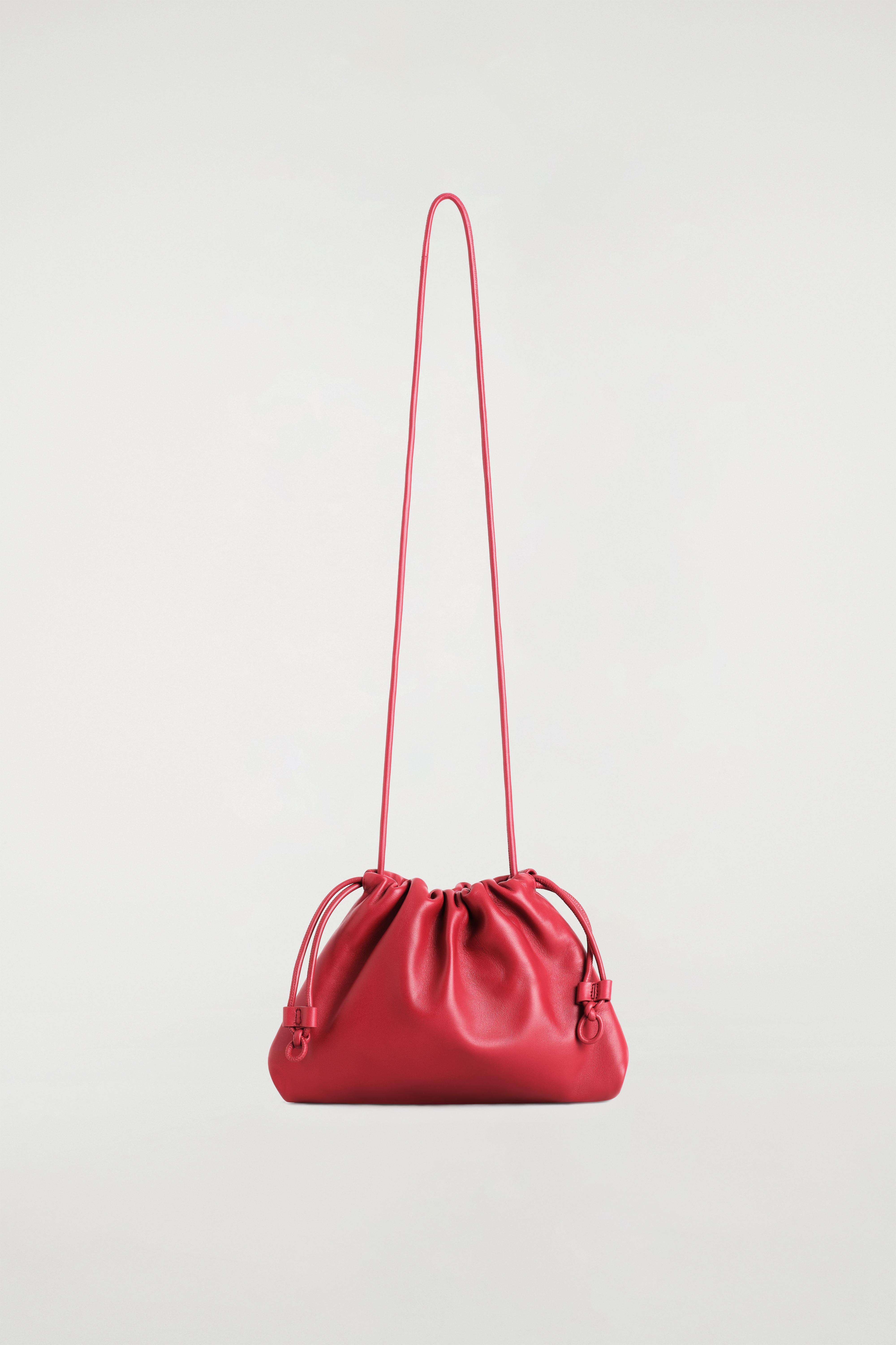 El bolso clutch Mini Cavatelli de piel roja se lleva al hombro por la modelo, que viste un abrigo largo negro. El bolso presenta una silueta fruncida, tipo bolsa, con finos detalles laterales de cordÃ³n ajustable y una correa de hombro a juego de piel.