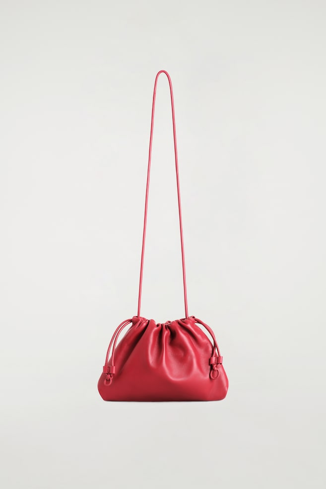 Le mini sac clutch Cavatelli en cuir rouge est présenté de face, mettant en valeur une silhouette souple et froncée avec une fermeture supérieure de style cordon coulissant. Il est suspendu à une longue bandoulière fine assortie en cuir, agrémentée de détails de cordon coulissant en cuir noués en boucle de chaque côté. - 1