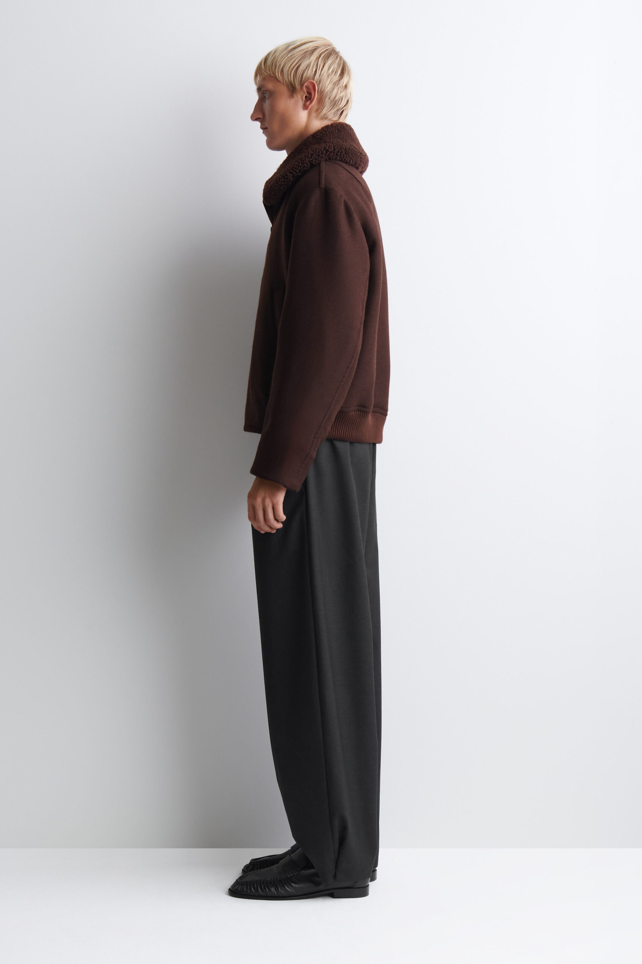 A model is wearing a dark brown jacket featuring a plush, textured shearling collar, structured sleeves, and a ribbed knit hem, shown in left-facing profile. The jacket is styled with loose, wide-leg dark gray trousers and black leather loafers. A model is wearing a dark brown jacket featuring a plush, textured shearling collar, structured sleeves, and a ribbed knit hem, shown in left-facing profile. The jacket is styled with loose, wide-leg dark gray trousers and black leather loafers.