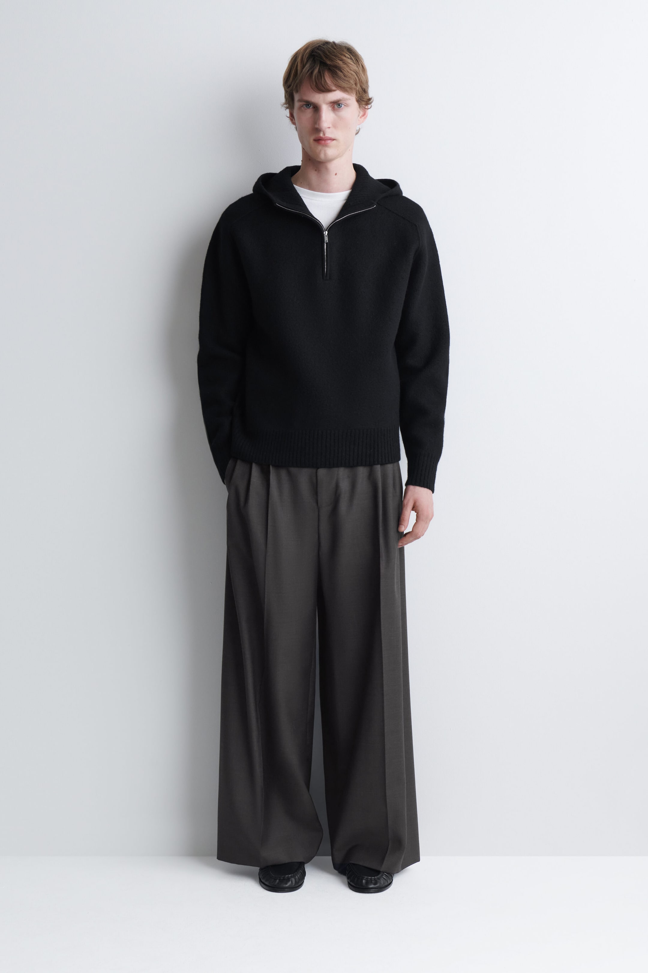 A black merino wool half-zip hoodie is worn by the model, featuring a silver-tone metal zip, a hood, and ribbed hem and cuffs. The hoodie is layered over a white crew-neck T-shirt and paired with dark grey wide-leg trousers. A black merino wool half-zip hoodie is worn by the model, featuring a silver-tone metal zip, a hood, and ribbed hem and cuffs. The hoodie is layered over a white crew-neck T-shirt and paired with dark grey wide-leg trousers.