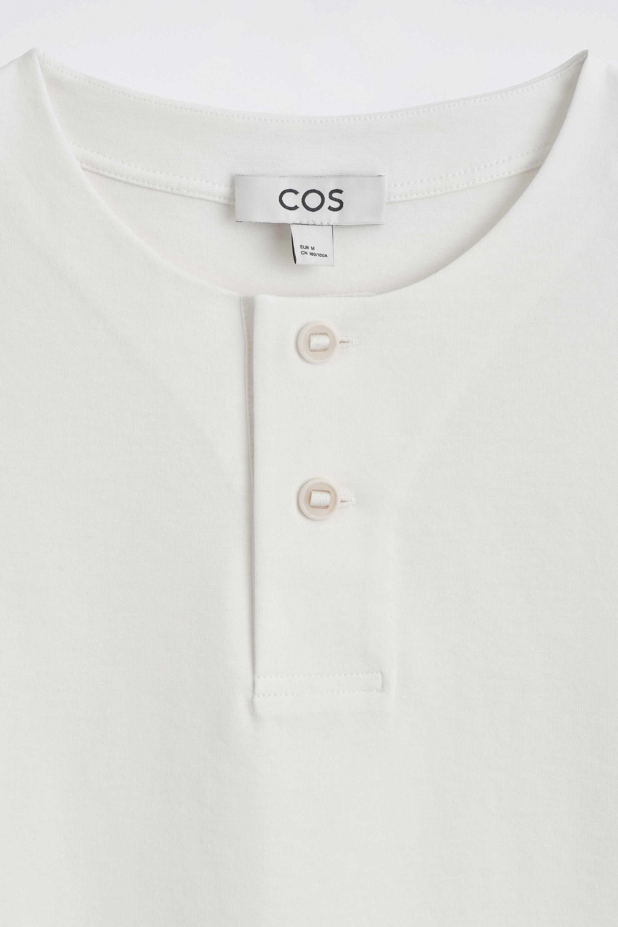 Le t-shirt Henley surdimensionné en jersey de coton est présenté en vue de face rapprochée, mettant en valeur son col rond et sa patte de boutonnage à deux boutons. Le vêtement blanc cassé présente une demi-patte de boutonnage cousue avec deux boutons beige pâle, ainsi qu’une étiquette de marque cousue à l’intérieur du col arrière.