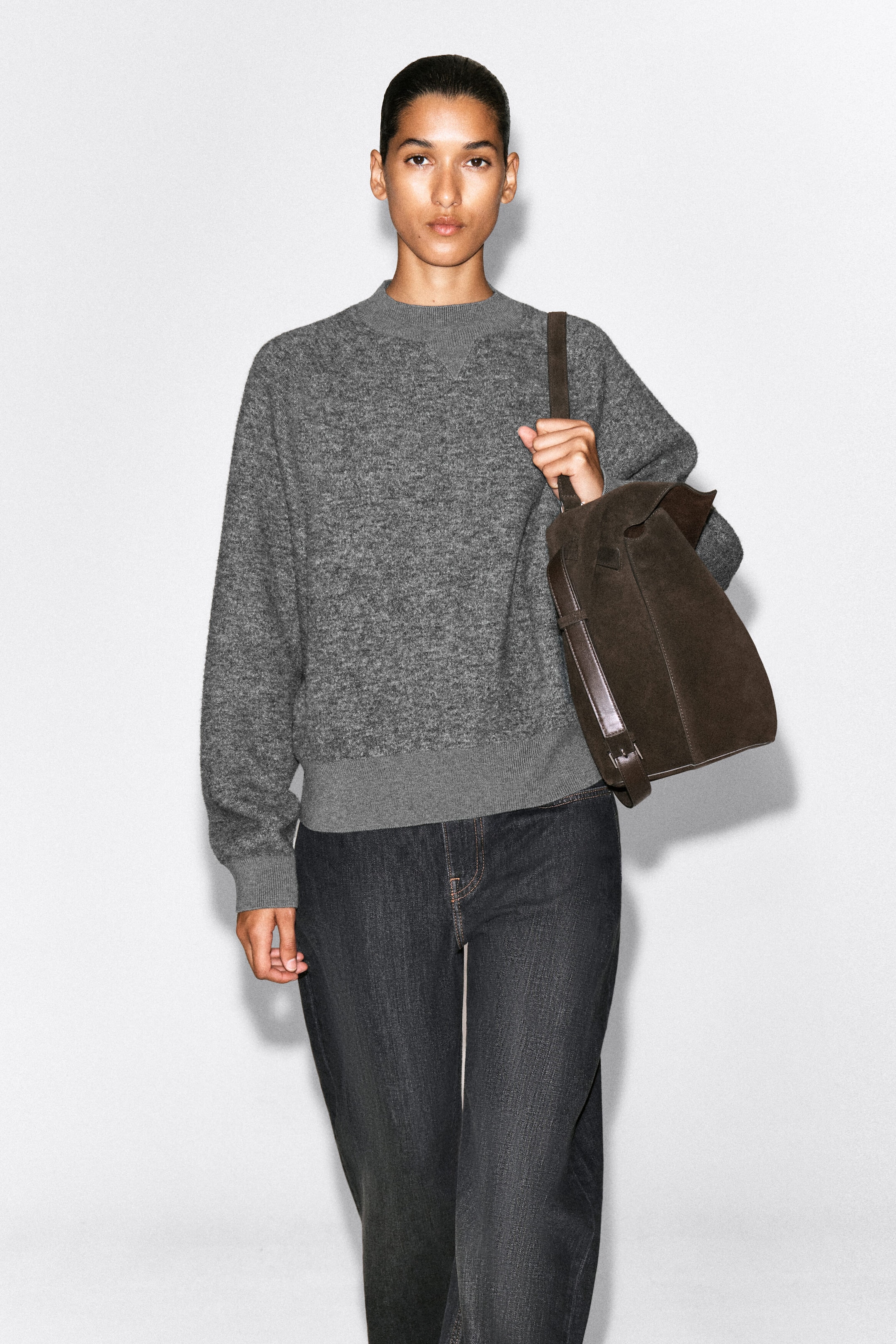 A gray textured knit sweater is shown from the front, featuring a crewneck collar with a triangular V-stitch detail, ribbed cuffs, and a wide ribbed hem. The relaxed-fit sweater is styled with dark gray denim jeans and a brown suede shoulder bag. A gray textured knit sweater is shown from the front, featuring a crewneck collar with a triangular V-stitch detail, ribbed cuffs, and a wide ribbed hem. The relaxed-fit sweater is styled with dark gray denim jeans and a brown suede shoulder bag.