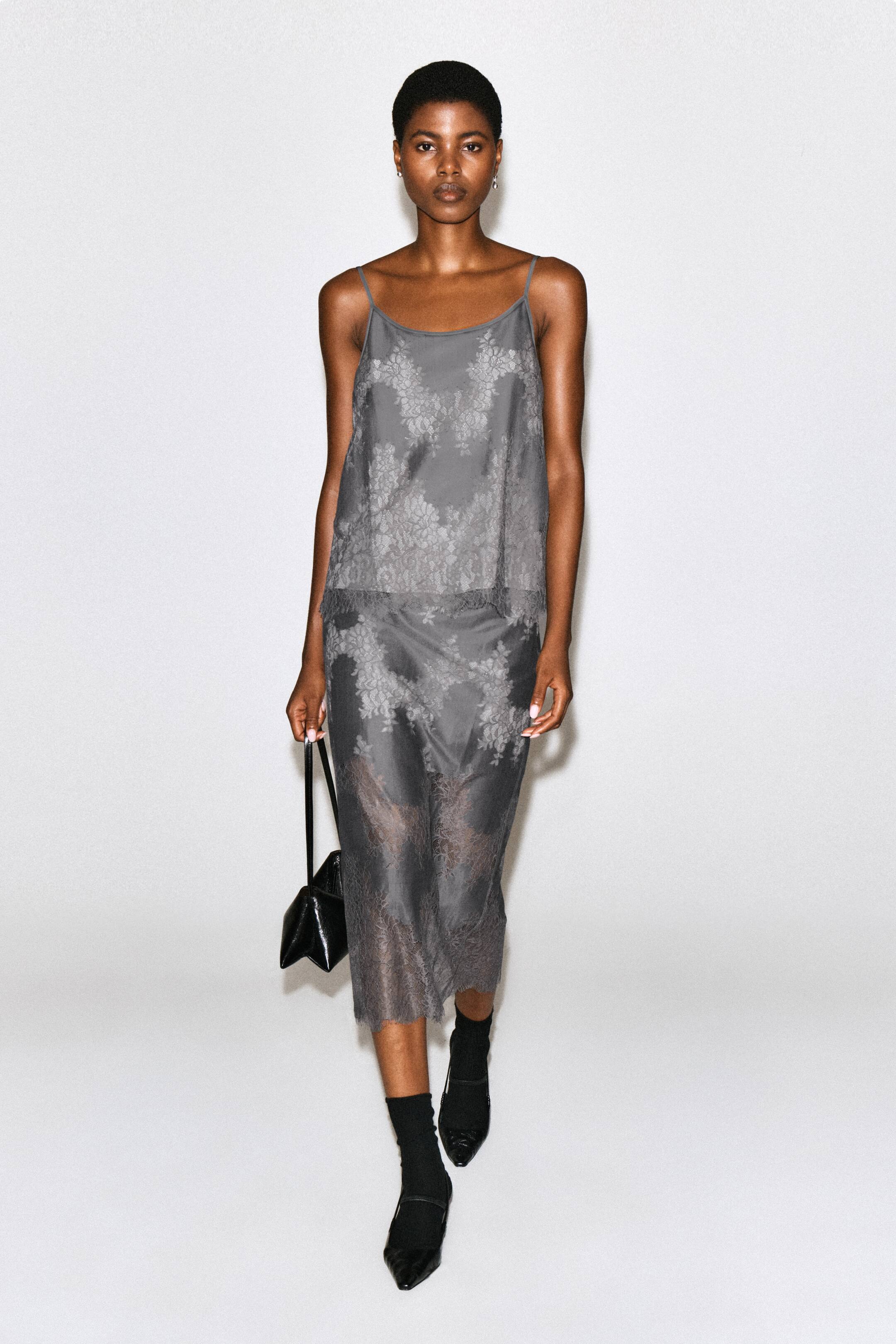 View larger image: A grey lace-overlay midi skirt featuring a floral pattern and a sheer, scalloped hem is being worn by the model. The skirt is styled with a matching lace camisole top, black socks, pointed-toe shoes, and a black handbag being carried in one hand.
