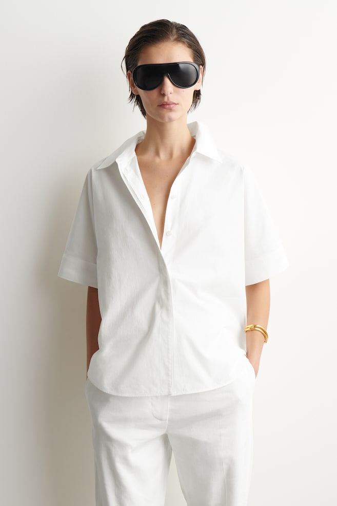 Se lleva puesta una camisa blanca de manga corta con botones al frente, que presenta un cuello en punta, puños doblados y un ajuste holgado, combinada con pantalones blancos a juego de corte holgado. - 1