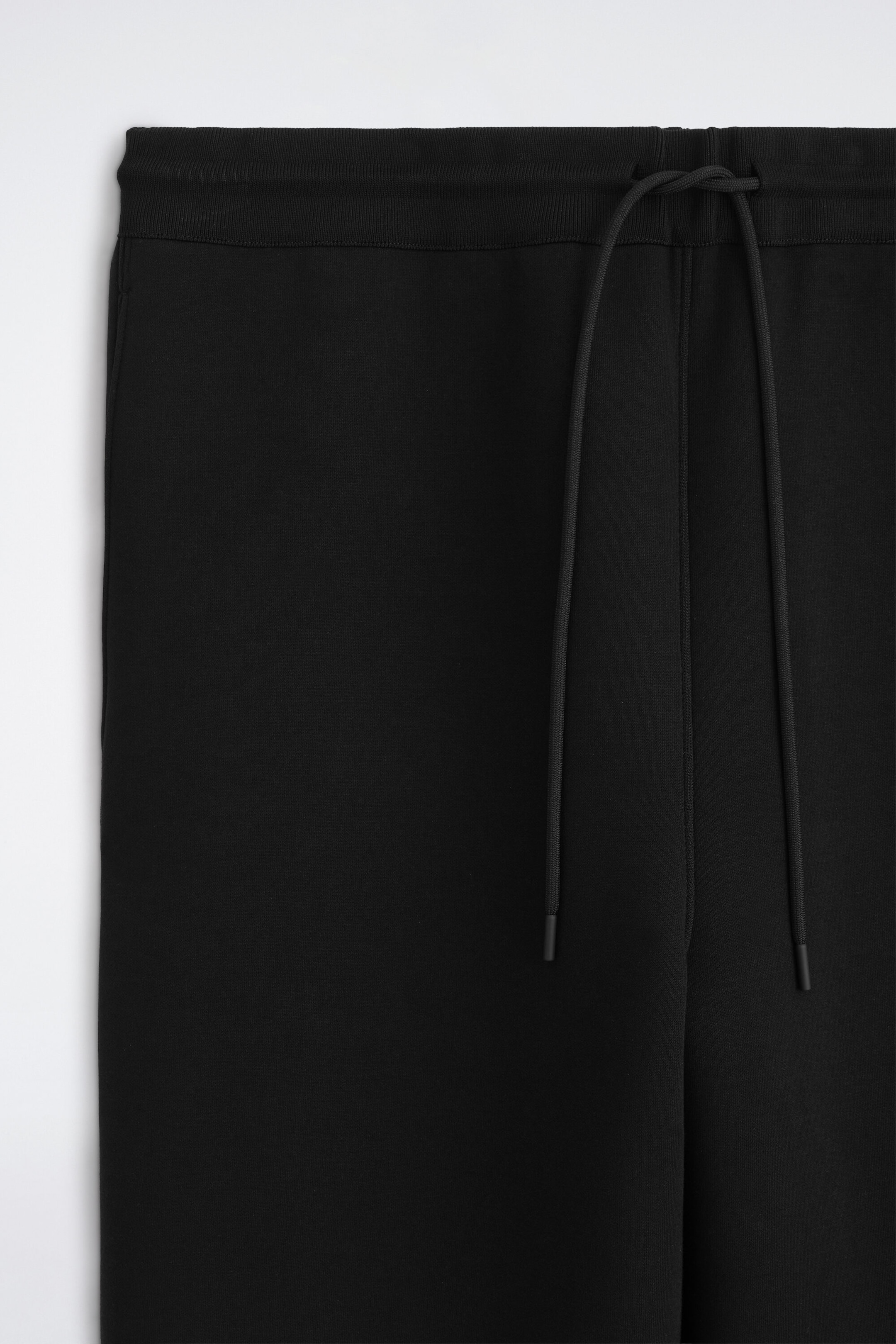 Black sweatpants are shown from the front, featuring a ribbed elastic waistband with a knotted black drawstring. The two long drawstrings with capped ends hang down the front of the smooth, dark fabric.