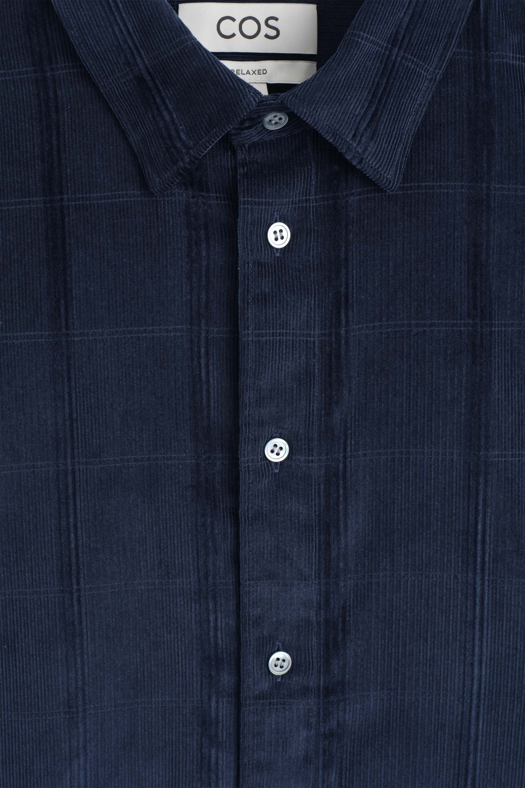 The Relaxed Corduroy Shirt is shown in a close-up front view, highlighting its dark navy-blue corduroy fabric with a subtle grid pattern. The shirt features a classic point collar, a button-down placket with light grey buttons, and a visible brand label at the inner neckline. The Relaxed Corduroy Shirt is shown in a close-up front view, highlighting its dark navy-blue corduroy fabric with a subtle grid pattern. The shirt features a classic point collar, a button-down placket with light grey buttons, and a visible brand label at the inner neckline.