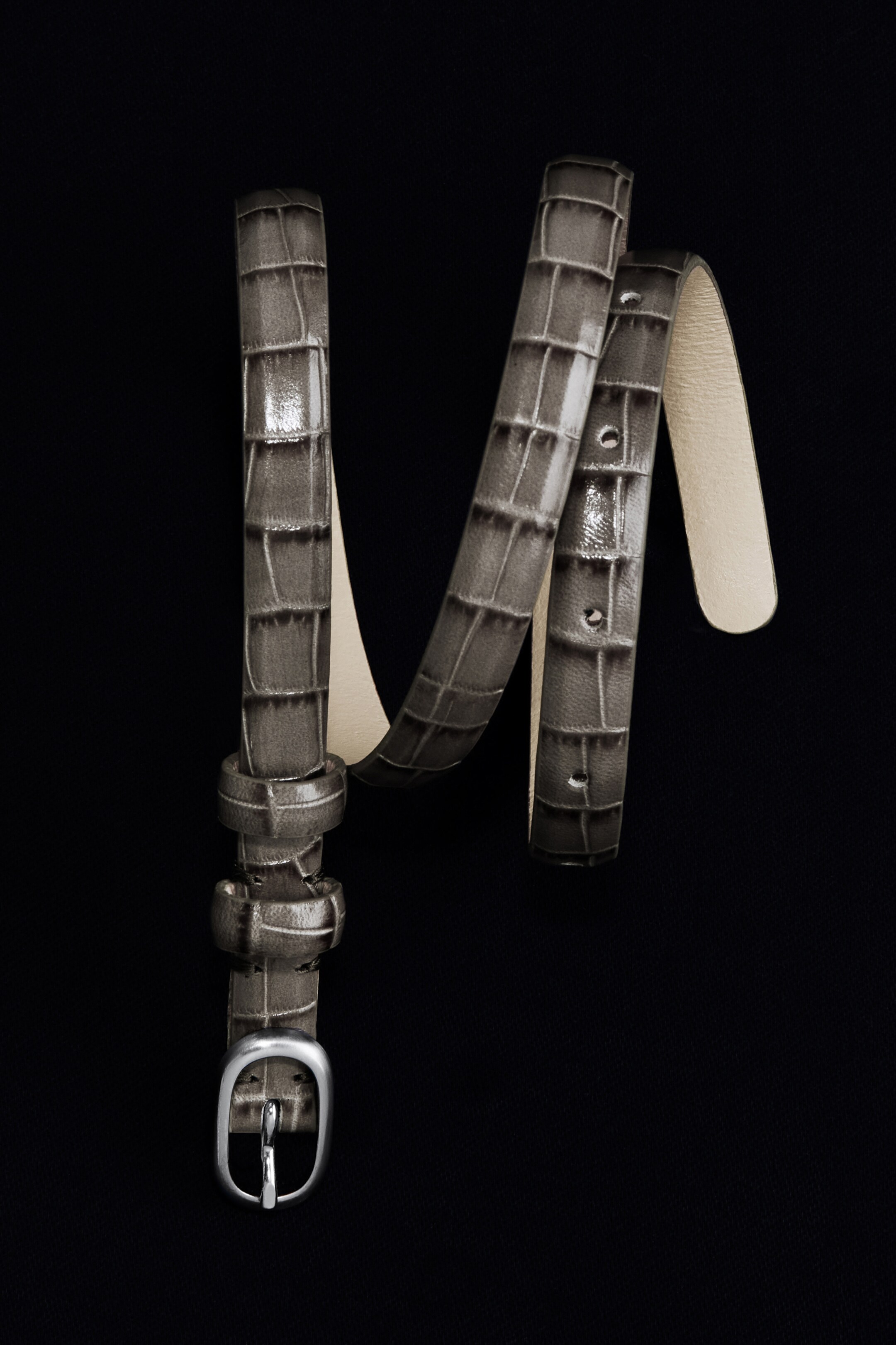 Größeres Bild anzeigen: Ein schlanker, taupe-grauer Ledergürtel windet sich und zeigt ein geprägtes Krokodilmuster. Er verfügt über eine skulpturale, silberfarbene ovale Schnalle und eine hellfarbene Unterseite.
