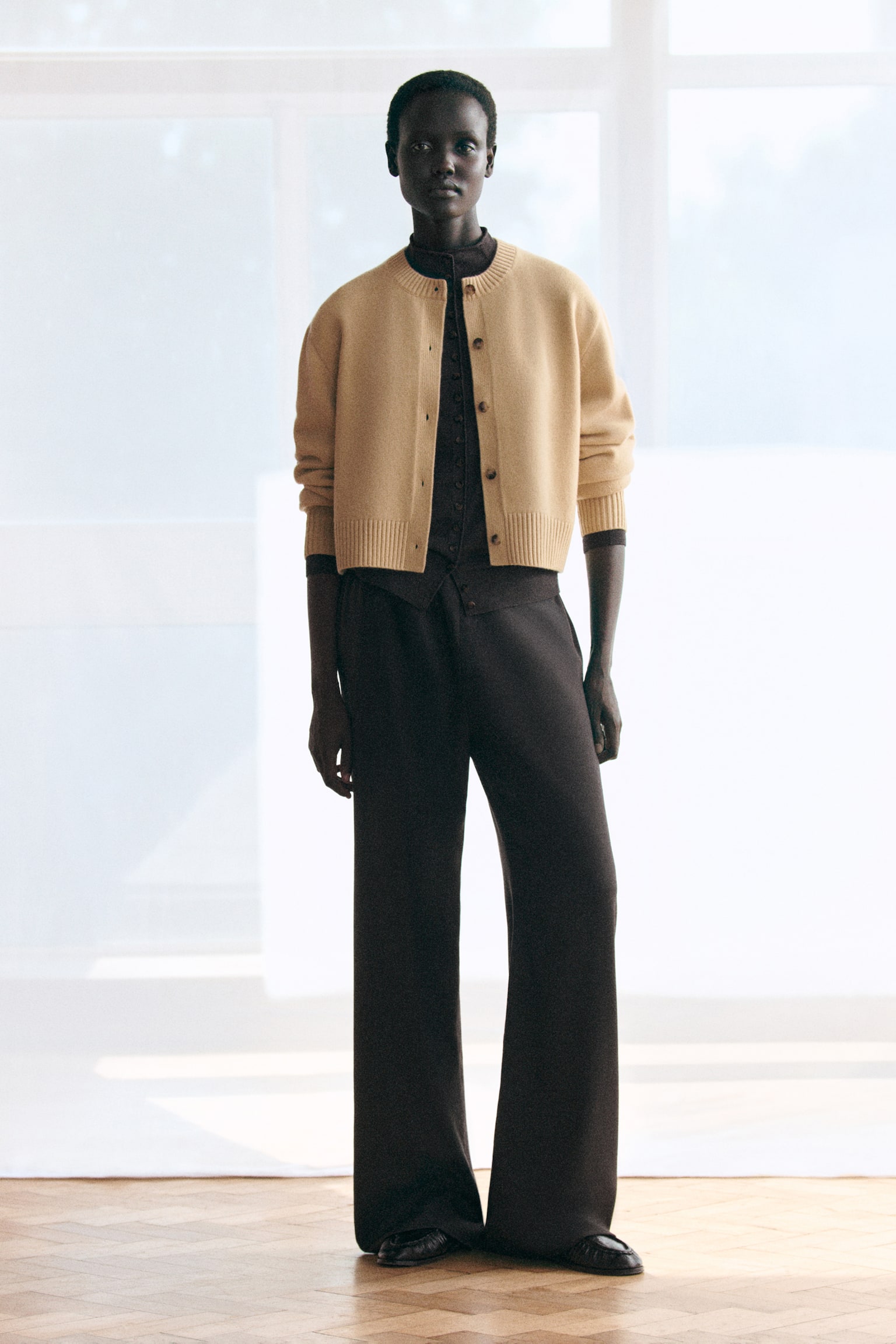 Een cropped, beige cardigan van merinowol met ronde hals en geribde afwerking wordt gedragen over een donkerbruine cardigan van merinowol met opstaande hals, beide van voren weergegeven om hun zachte texturen en relaxte pasvormen te benadrukken. De donkerbruine cardigan met opstaande hals is dichtgeknoopt en gecombineerd met bijpassende broek met wijde pijpen, wat zorgt voor een gecoördineerd, gedrapeerd silhouet. - 1